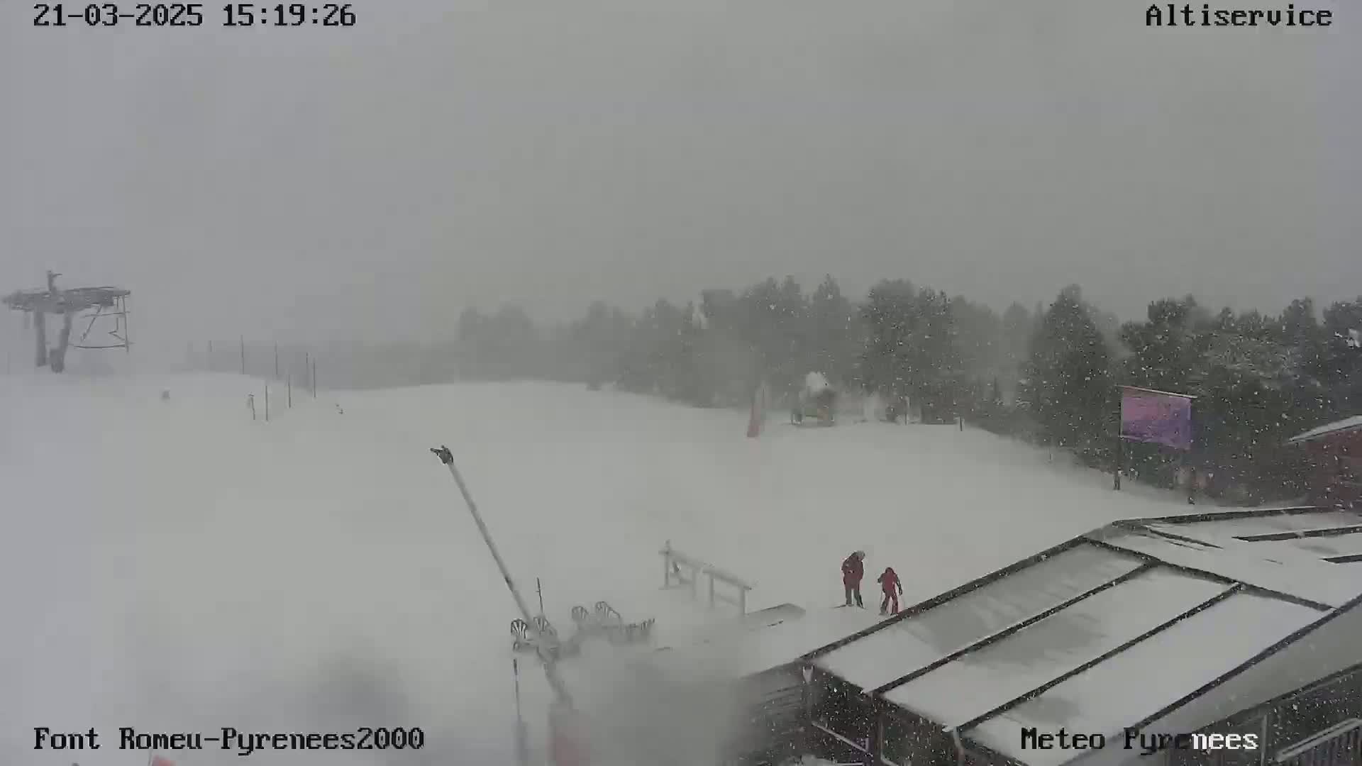 A snowy scene at a ski resort shows a heavy snowfall obscuring the view of trees and buildings, with a few people visible near a ski lift.