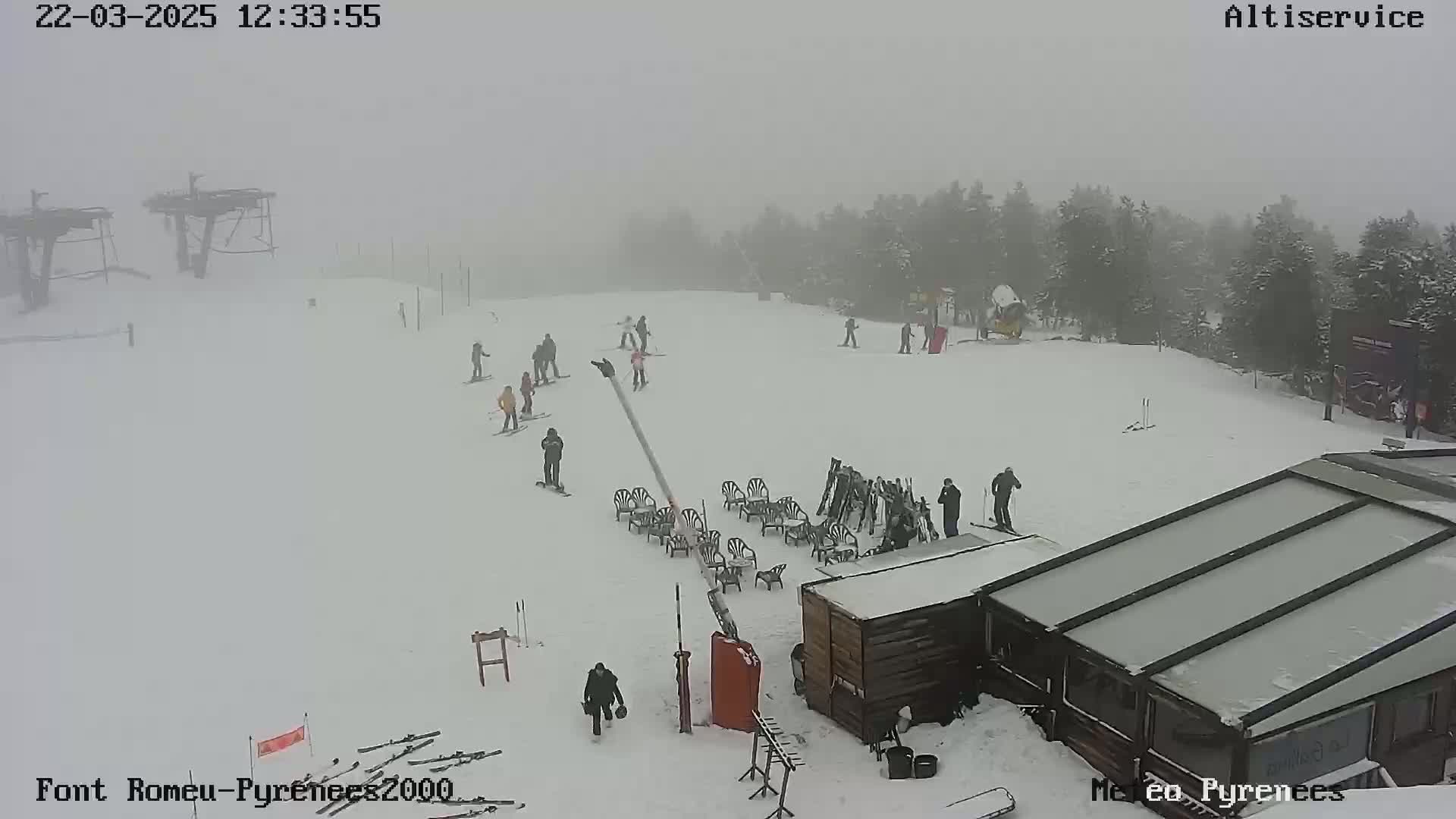 A snowy ski slope with several skiers and snowboarders visible in foggy conditions, near a building and ski lift.