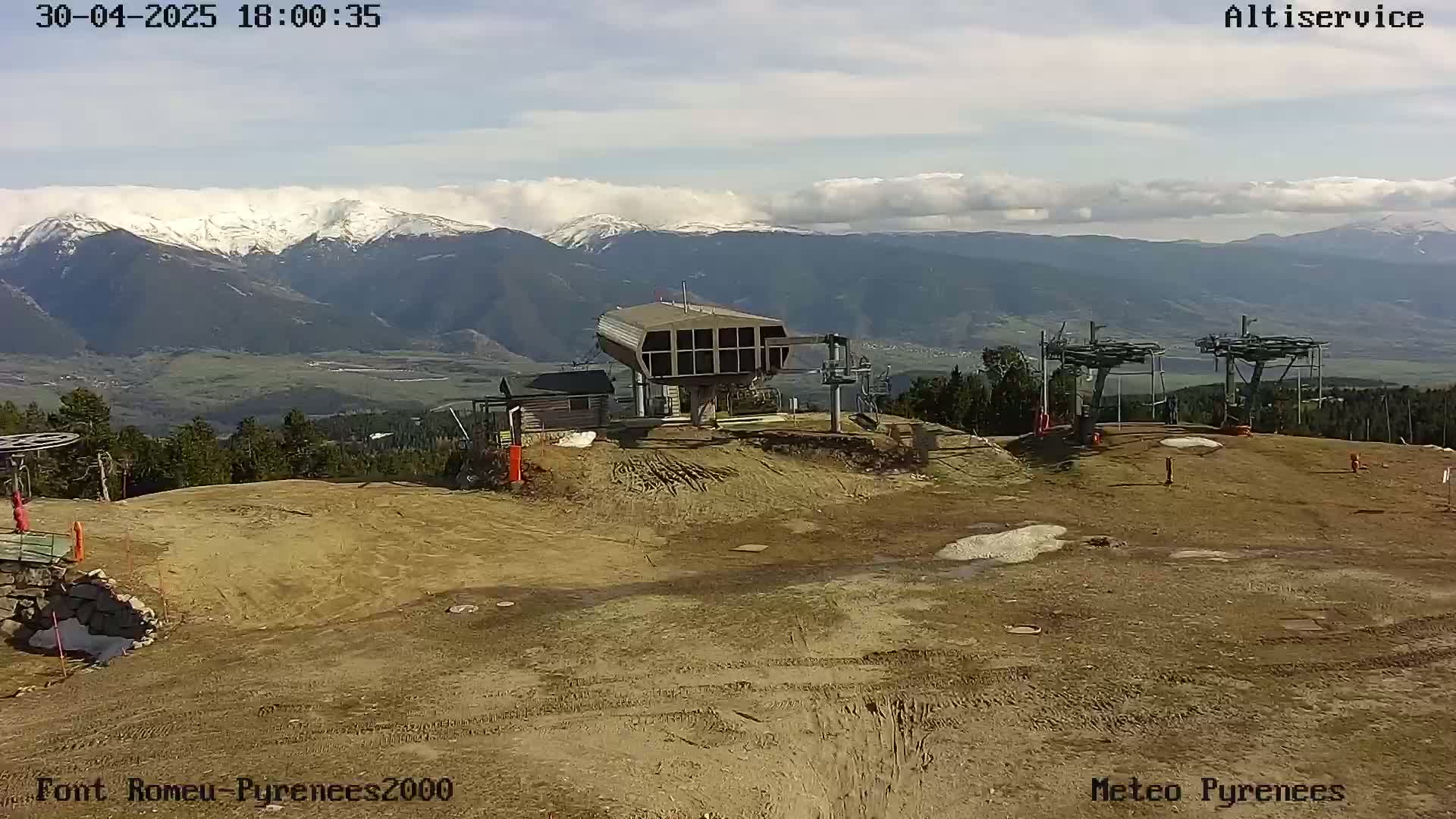 A mostly sunny day reveals a ski lift station and surrounding bare, brown land, with snow-capped mountains in the distance.