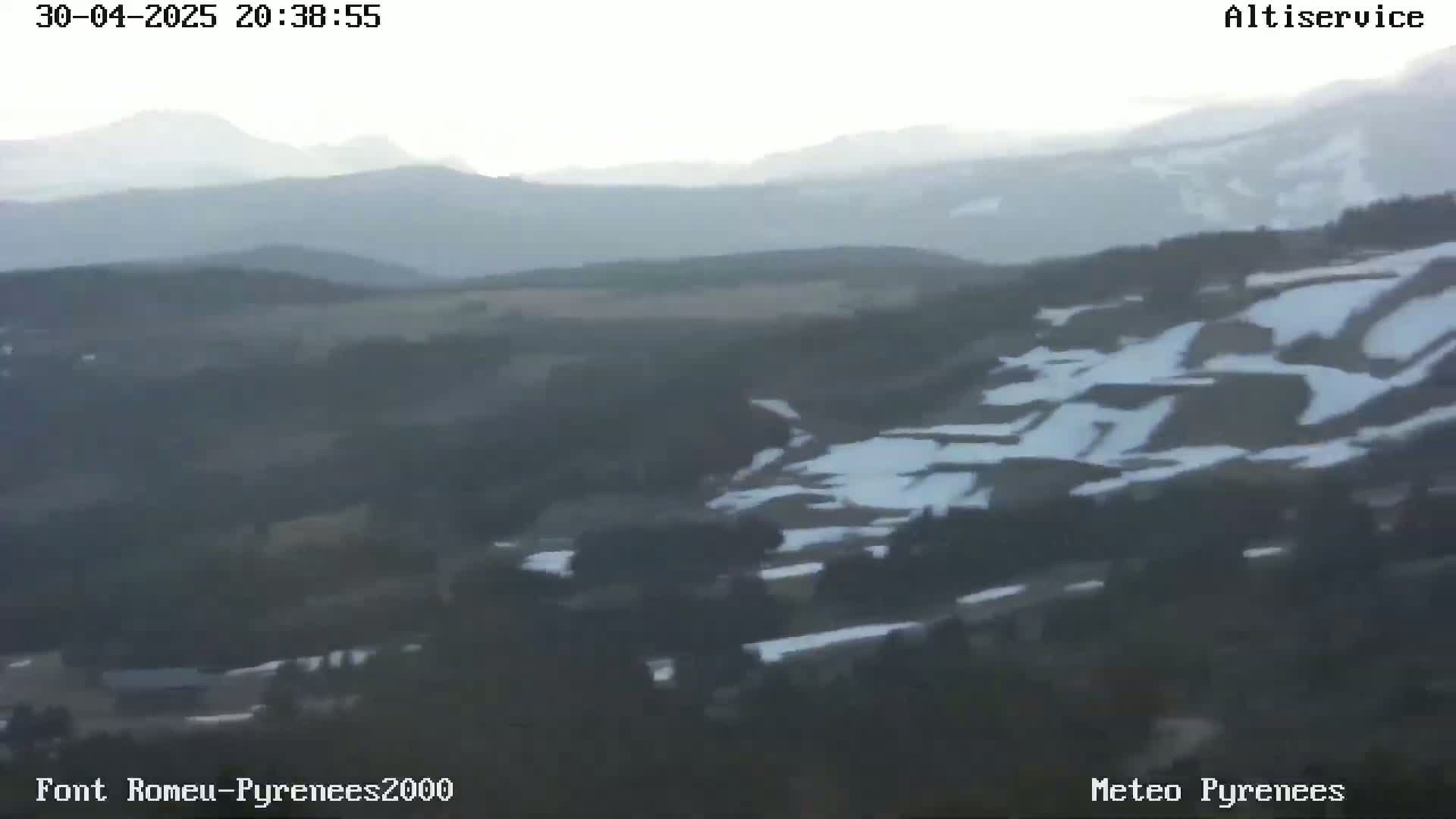 A hazy, long-range view of a mountainous landscape with patches of snow, showing a valley in the foreground and distant mountains under a somewhat clear sky.