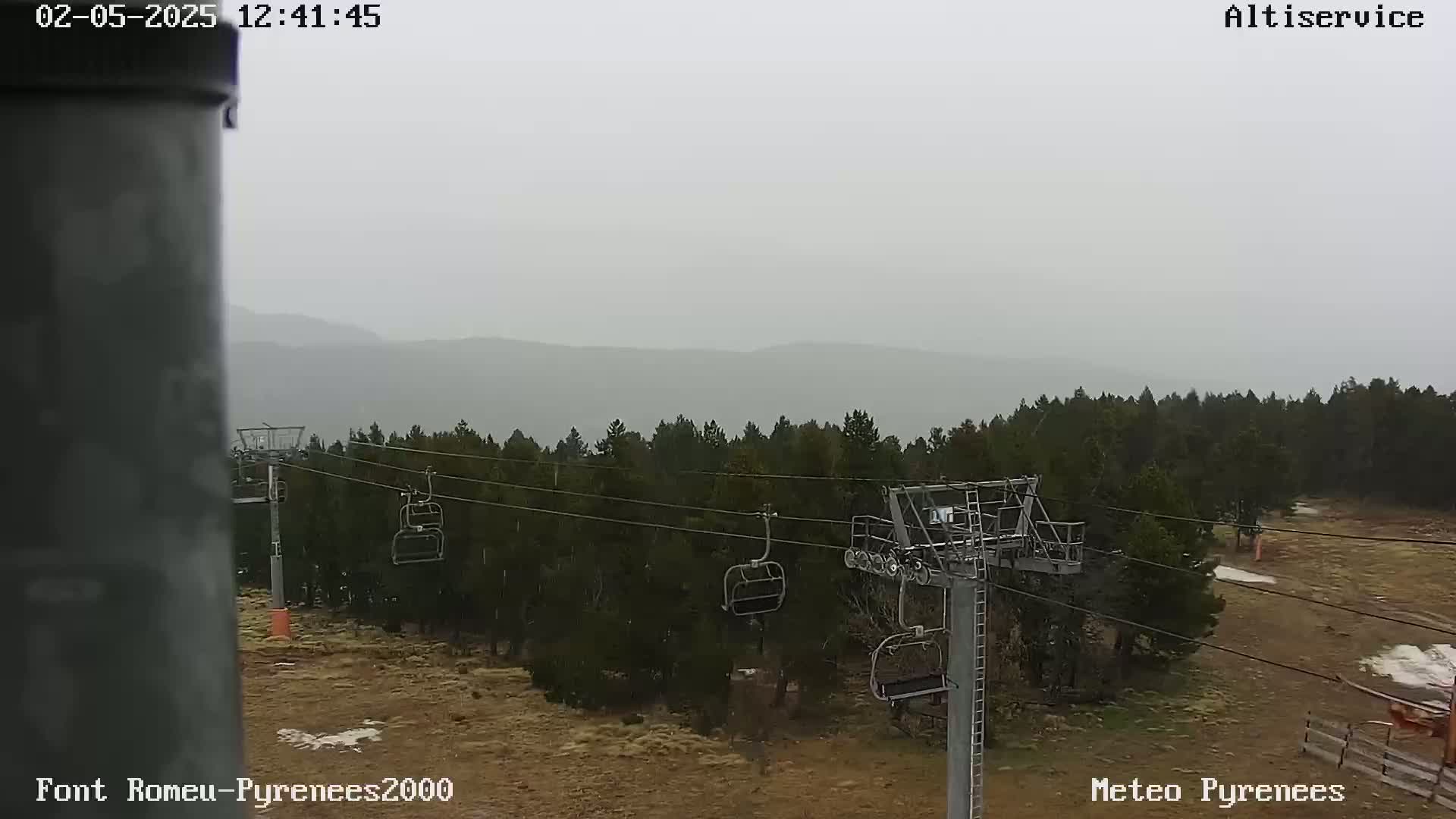 A chairlift stands in a snow-dusted field of brown grass and dark evergreens, with hazy mountains visible in the distance under a gray sky.