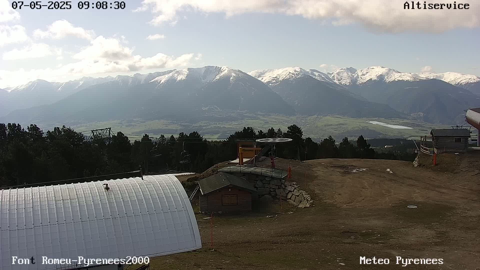 A mostly sunny view of snow-capped mountains in the distance, seen from a ski resort with a small building and a chairlift pulley visible in the foreground.