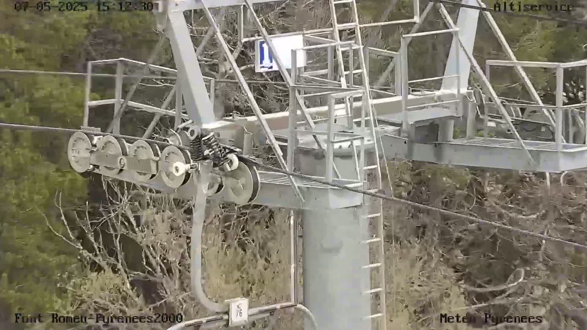 A close-up view of a chairlift's pulley system and support structure against a backdrop of bare, brown trees in overcast conditions.