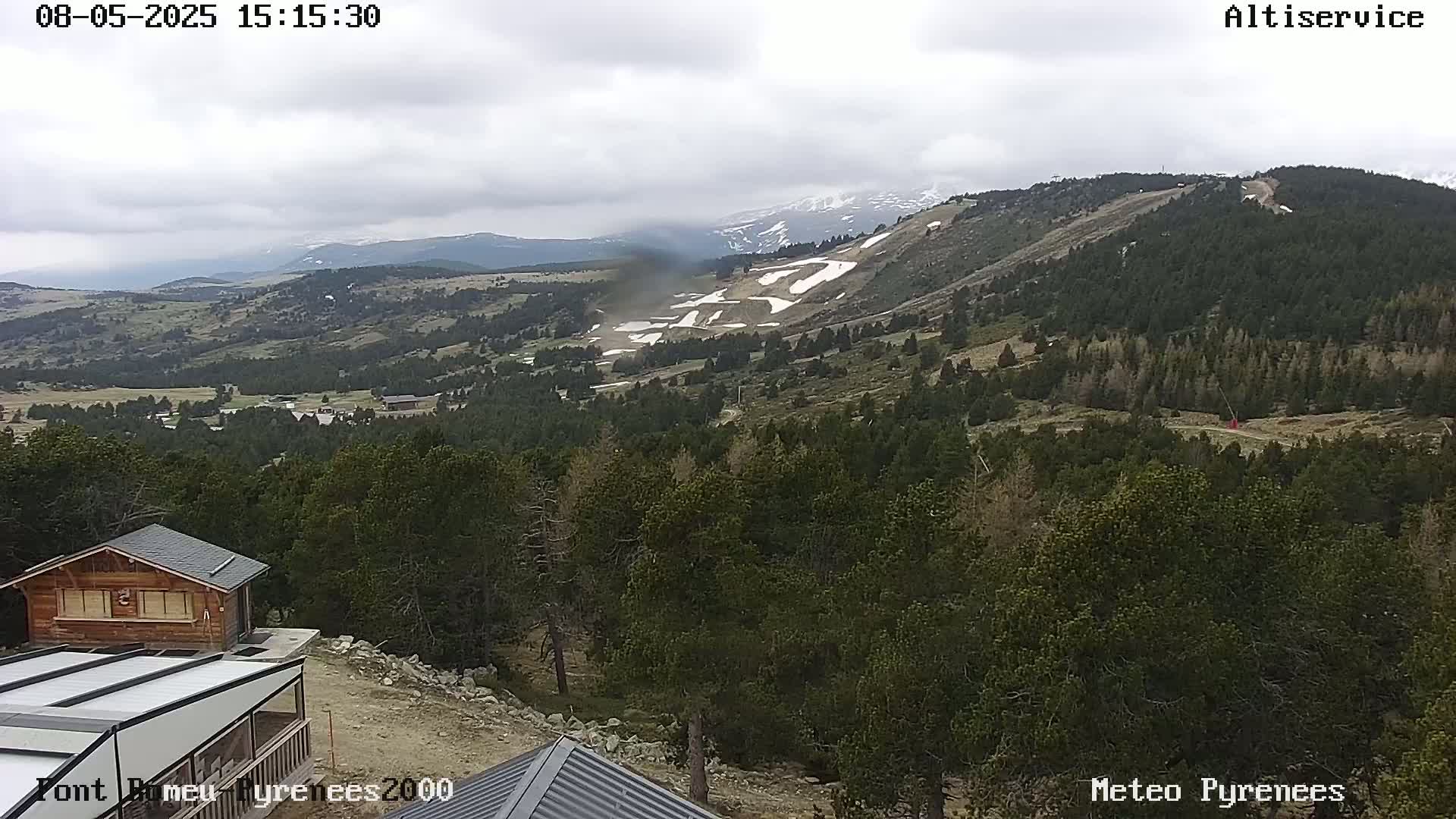 A mountain range with patches of snow under an overcast sky, viewed from a high vantage point overlooking a forest and small buildings.