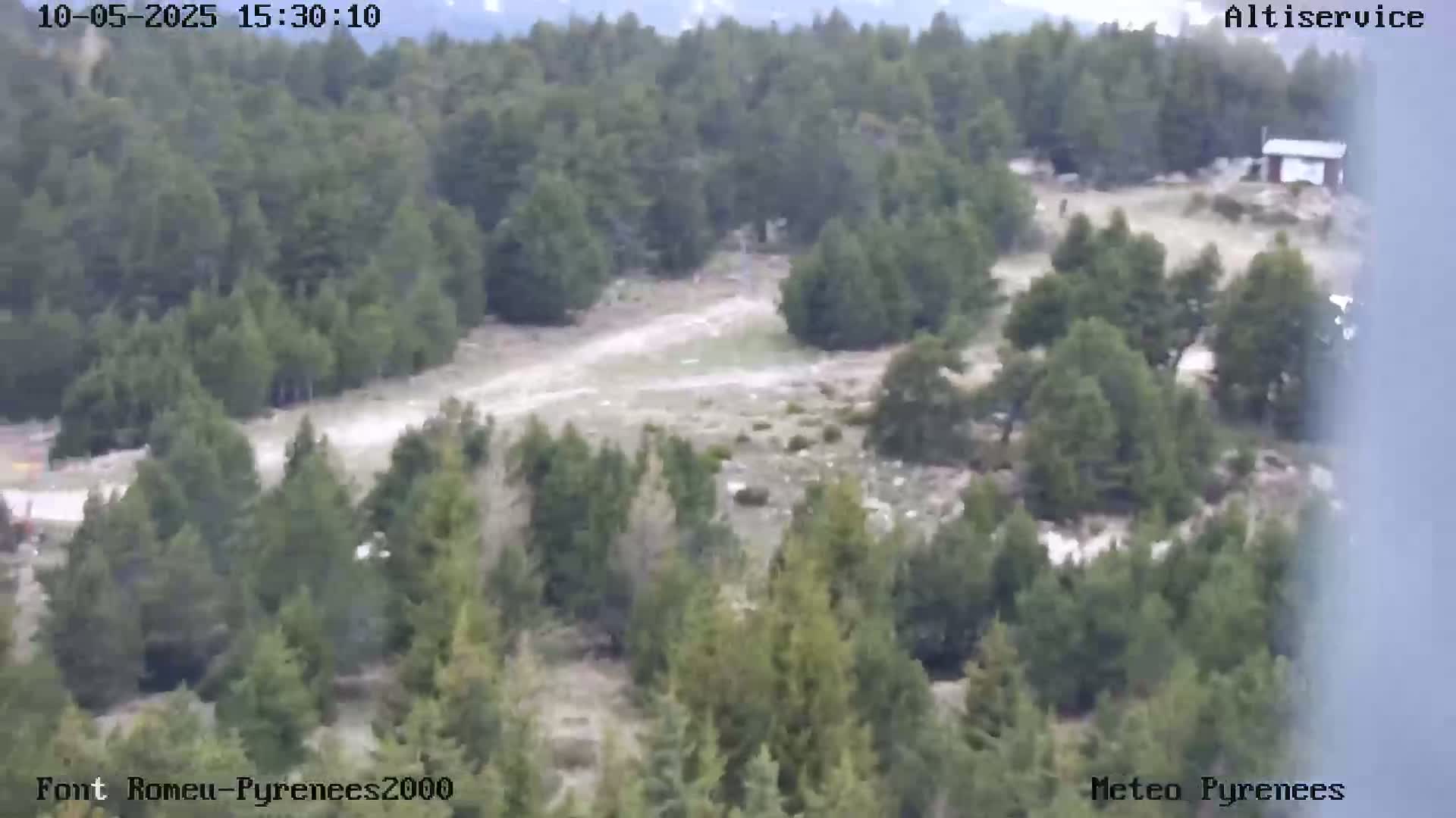 An aerial view shows a mostly clear, dry day over a mountainous area with sparse, light-colored ground cover and patches of evergreen trees.