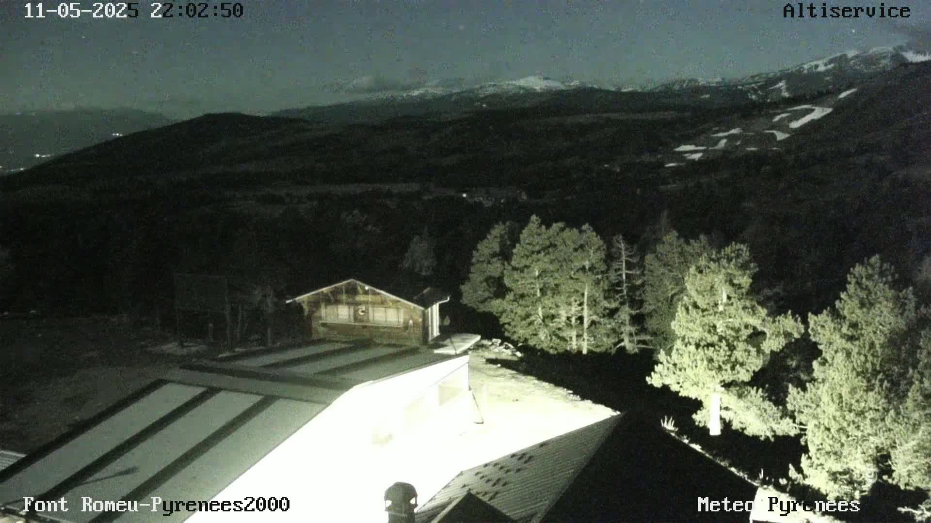 A nighttime view of snow-capped mountains in the distance, with illuminated buildings and evergreen trees in the foreground under a clear, dark sky.
