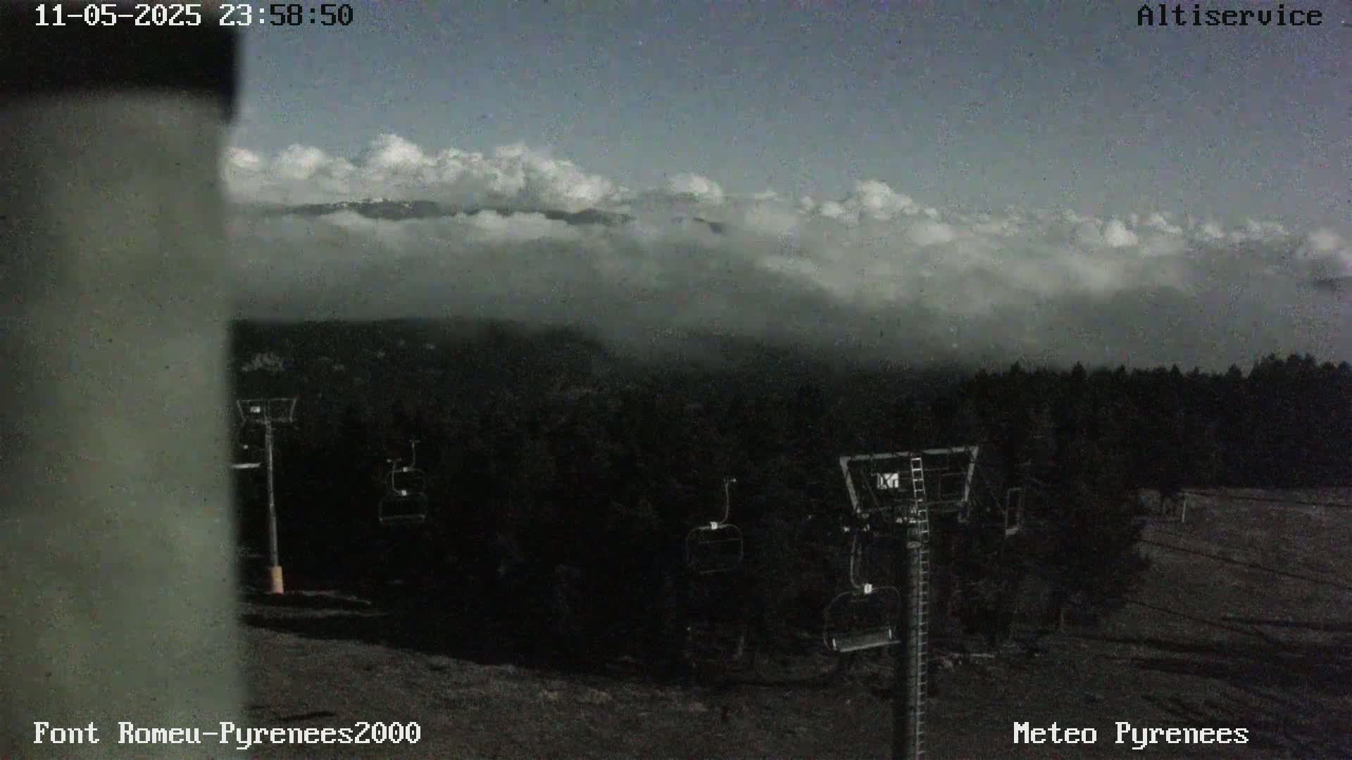 A cloudy, twilight scene shows several empty chairlifts in a ski resort with a dark forest and distant mountains partially obscured by low clouds.