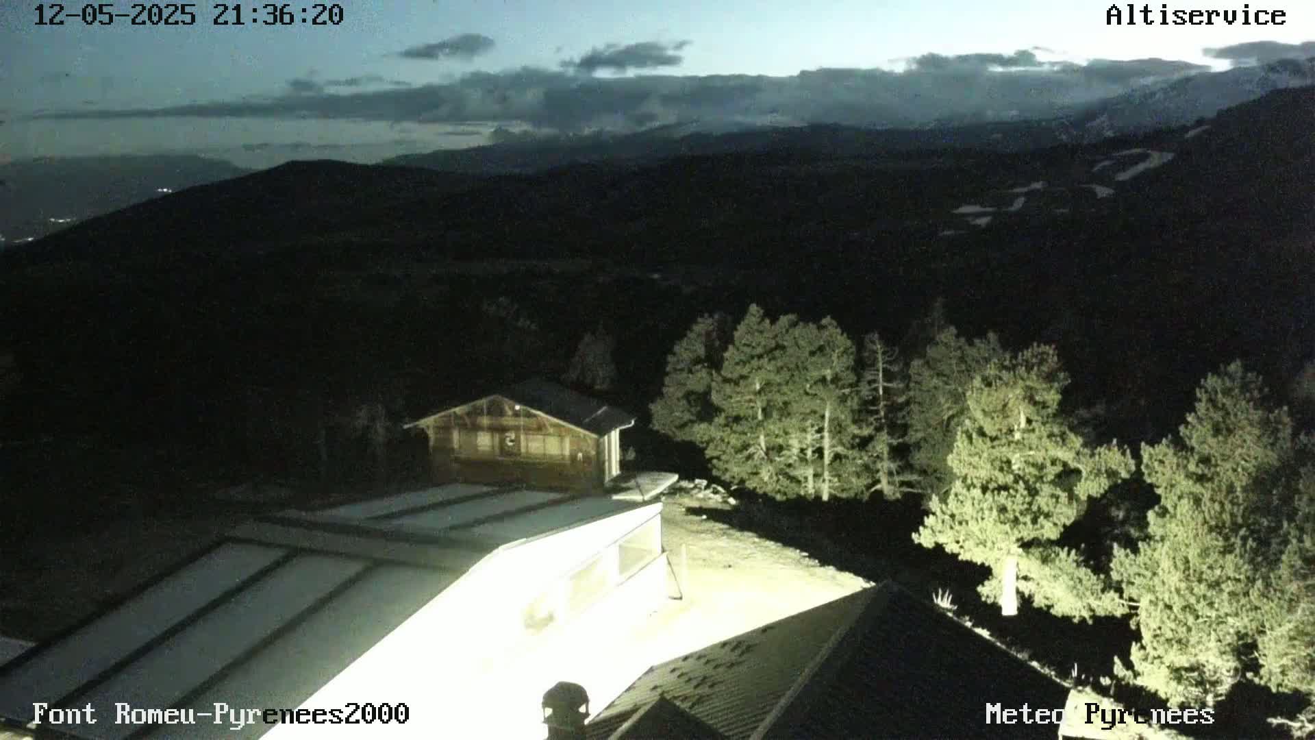 A nighttime view of a mountainous landscape under a partly cloudy sky shows several buildings nestled among dark evergreens, with distant lights visible in the valley below.