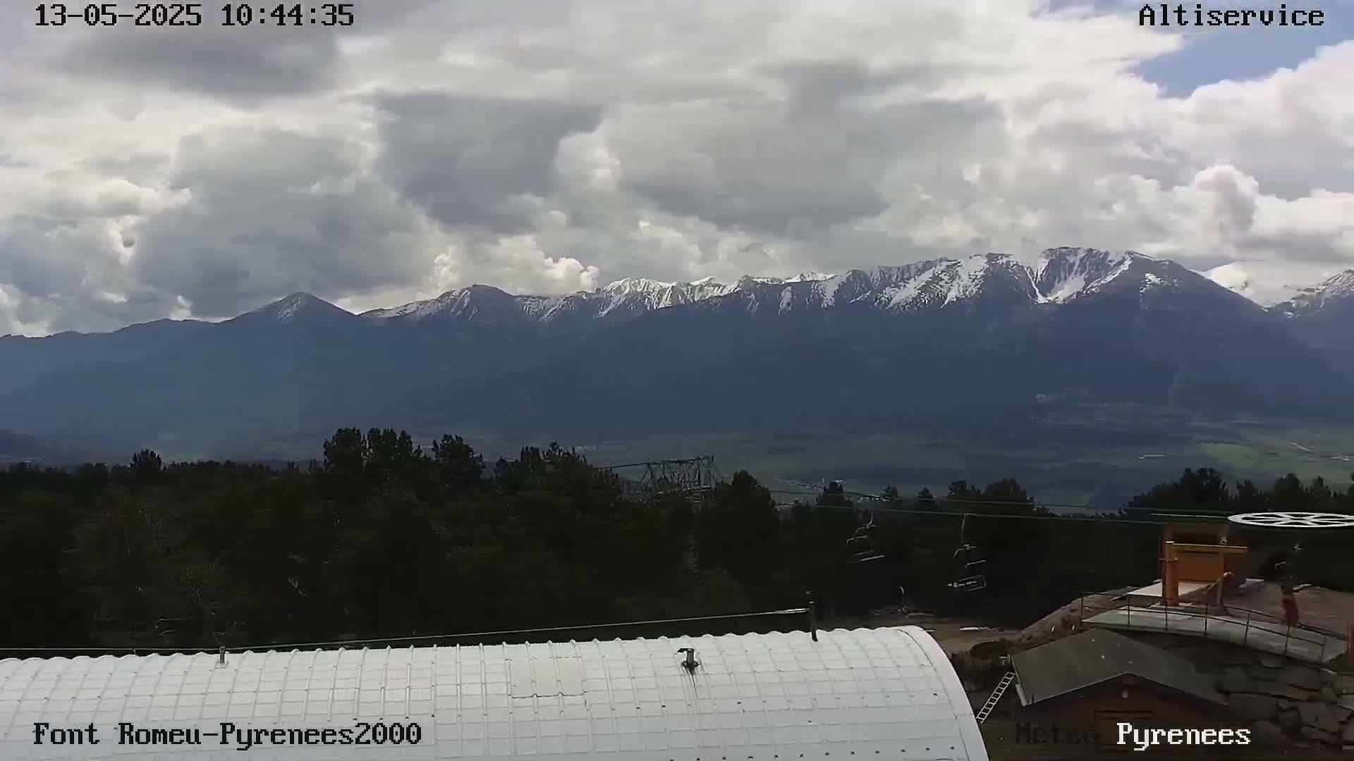 A view of snow-capped mountains under a partly cloudy sky, with a foreground of trees, a building's roof, and a ski lift.