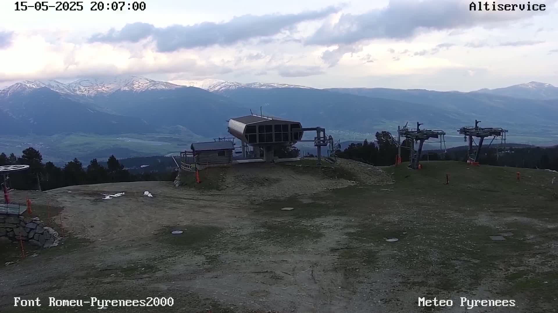A mostly cloudy, late evening view shows a ski lift station and chairlift supports on a barren, dusty mountaintop overlooking a valley and snow-capped mountains in the distance.