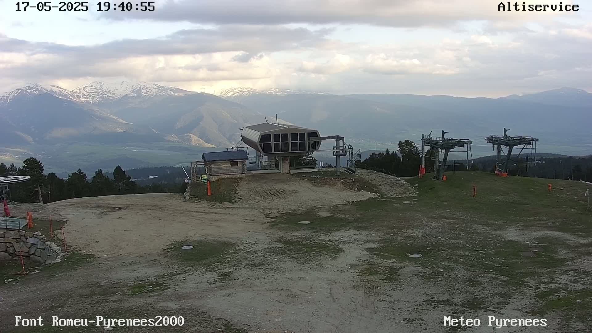 A ski lift station sits atop a hill overlooking a valley and snow-capped mountains under a cloudy sky.