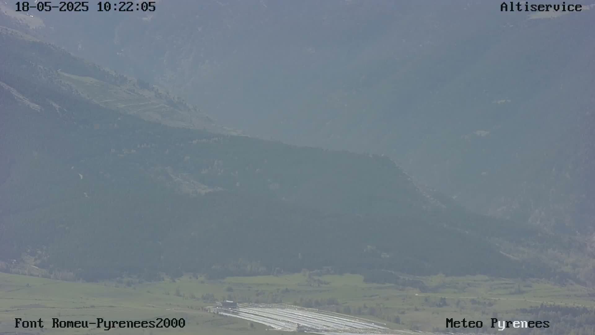 A hazy view shows a valley with a flat area and long, parallel lines, possibly a field, surrounded by mountains under a mostly clear sky.
