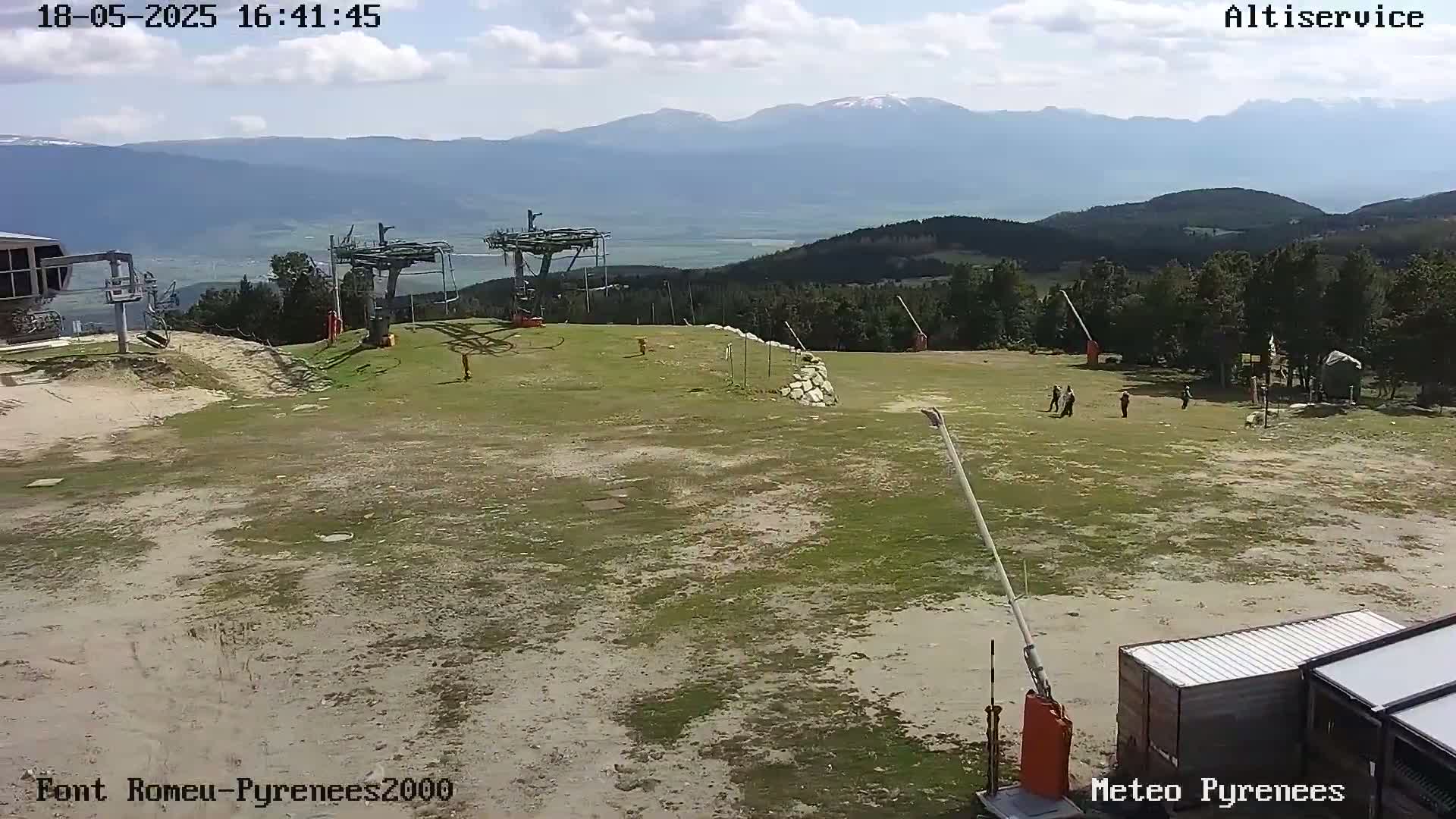 A mostly sunny day at a ski resort shows a grassy area with ski lifts, a few people, and distant mountains under a partly cloudy sky.