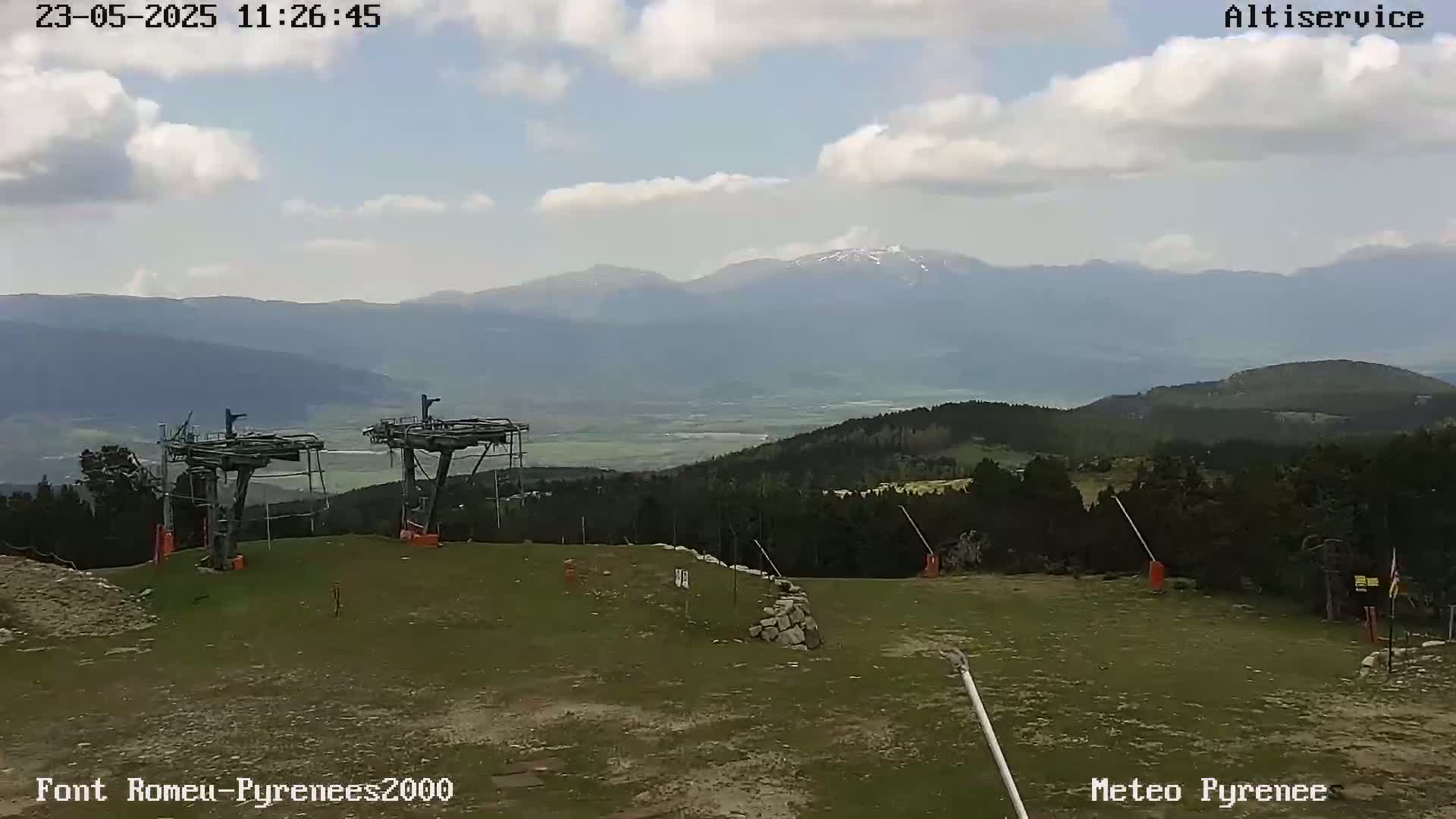 Two empty chairlift structures sit atop a grassy hill overlooking a valley and distant mountains under a partly cloudy sky.
