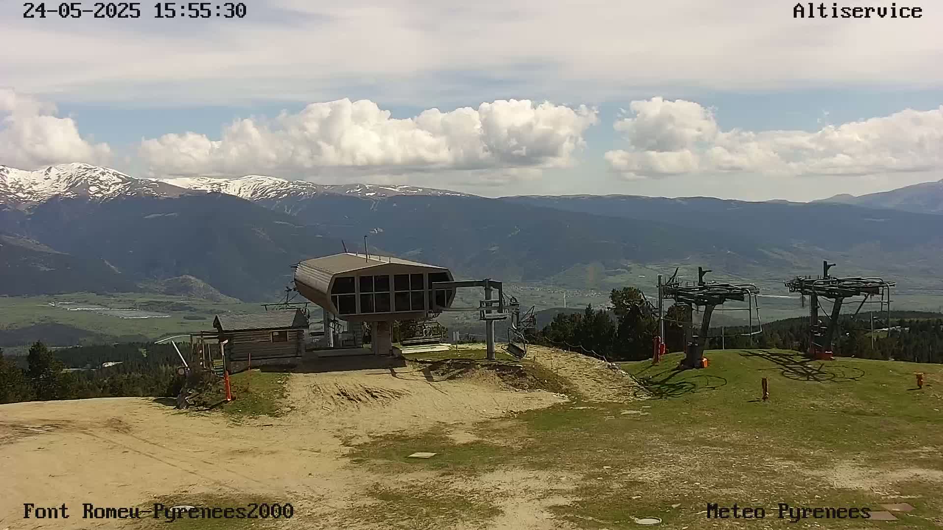 A partially cloudy sky overlooks a mountain range with snow-capped peaks, a ski lift station, and two chairlift towers in a grassy area.