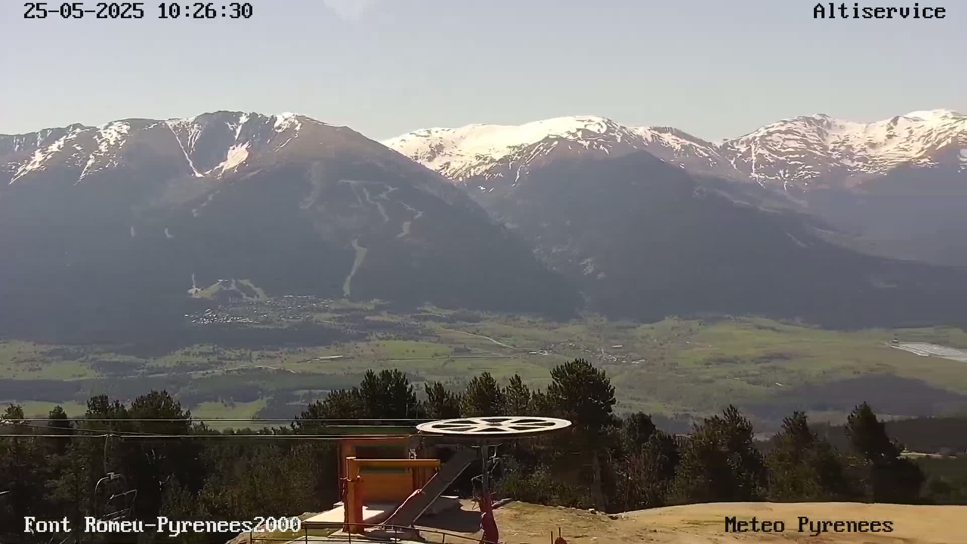 A mountaintop view shows snow-capped mountains in the distance, a valley of green fields and trees below, and a ski lift pulley in the foreground under a mostly clear sky.