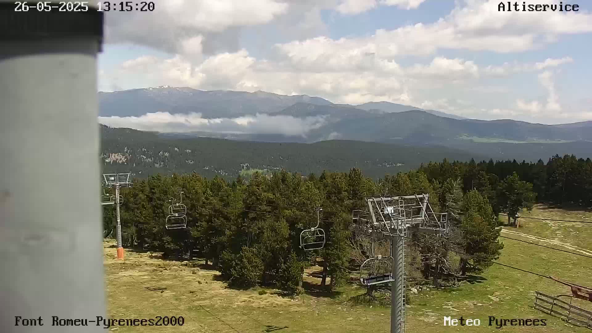 A partly cloudy day reveals a mountain range in the distance, with a ski lift and surrounding coniferous trees in the foreground.