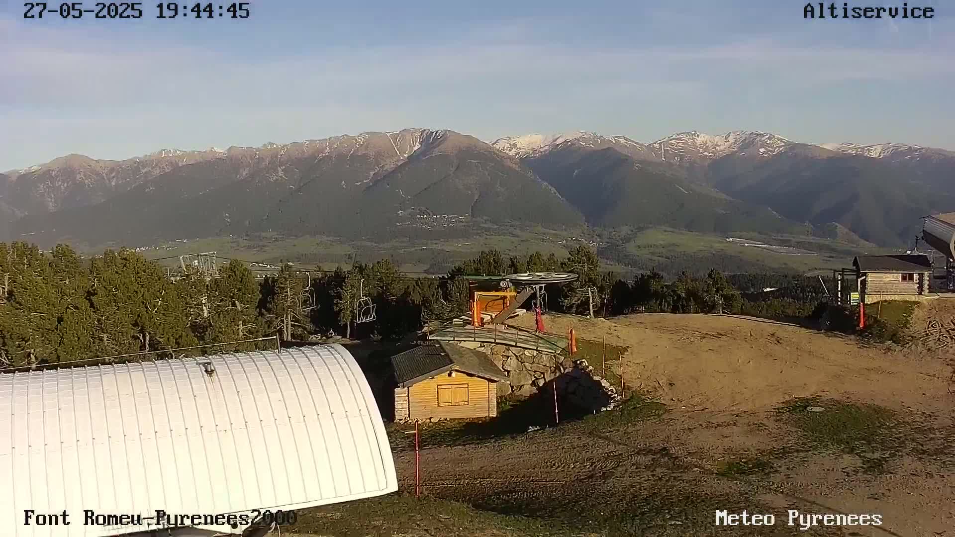 A mountaintop view shows a ski lift, a small wooden building, and a large white structure under a clear, sunny sky with snow-capped mountains in the background.