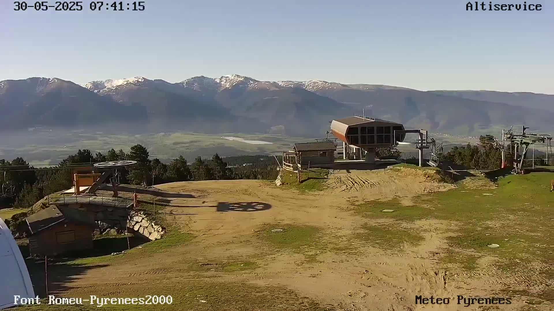 A ski lift station sits on a mostly barren, dusty hillside overlooking a valley and snow-capped mountains under a clear sky.
