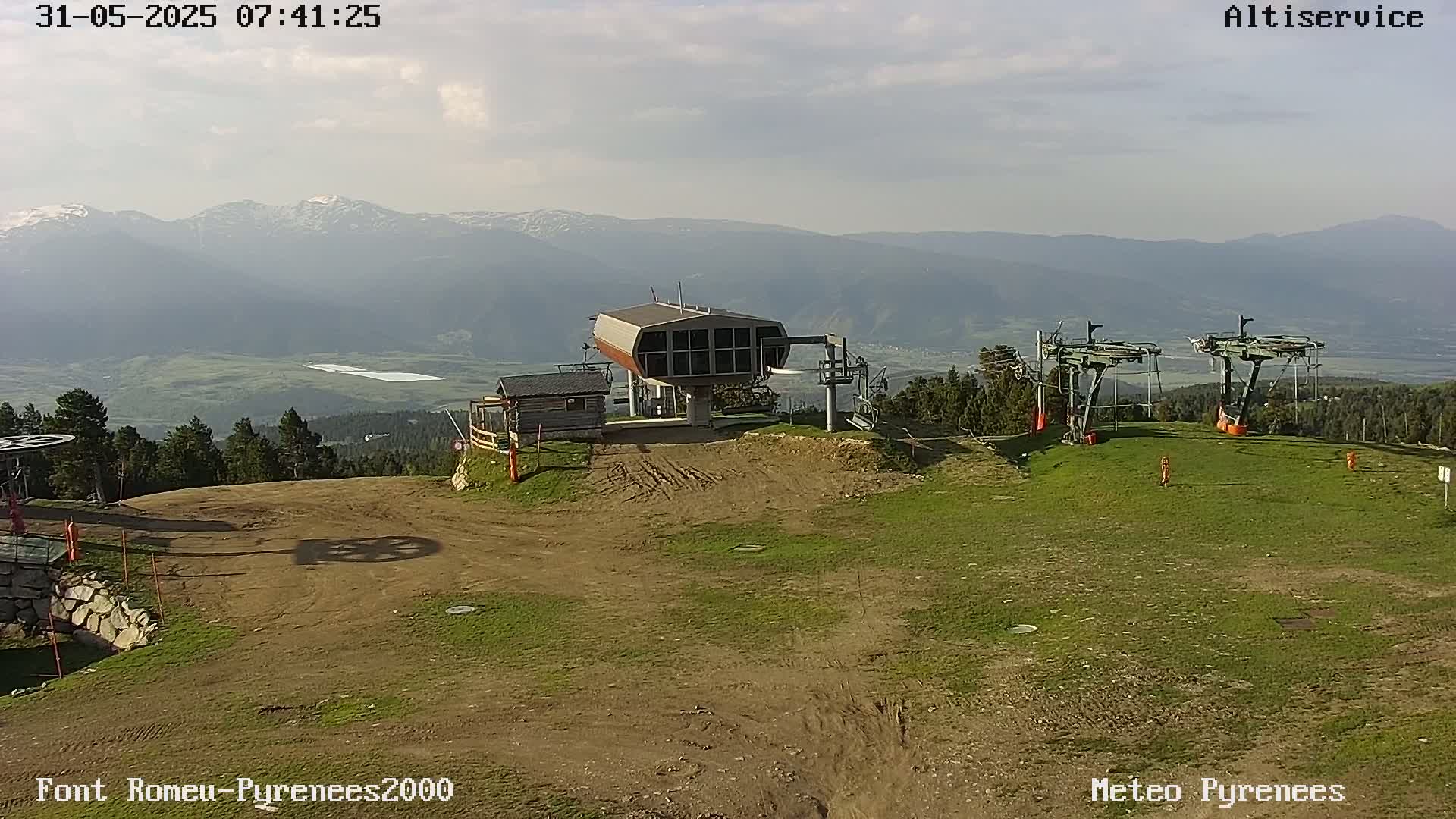 A chairlift station sits on a grassy hill overlooking a valley and distant mountains under a partly cloudy sky.