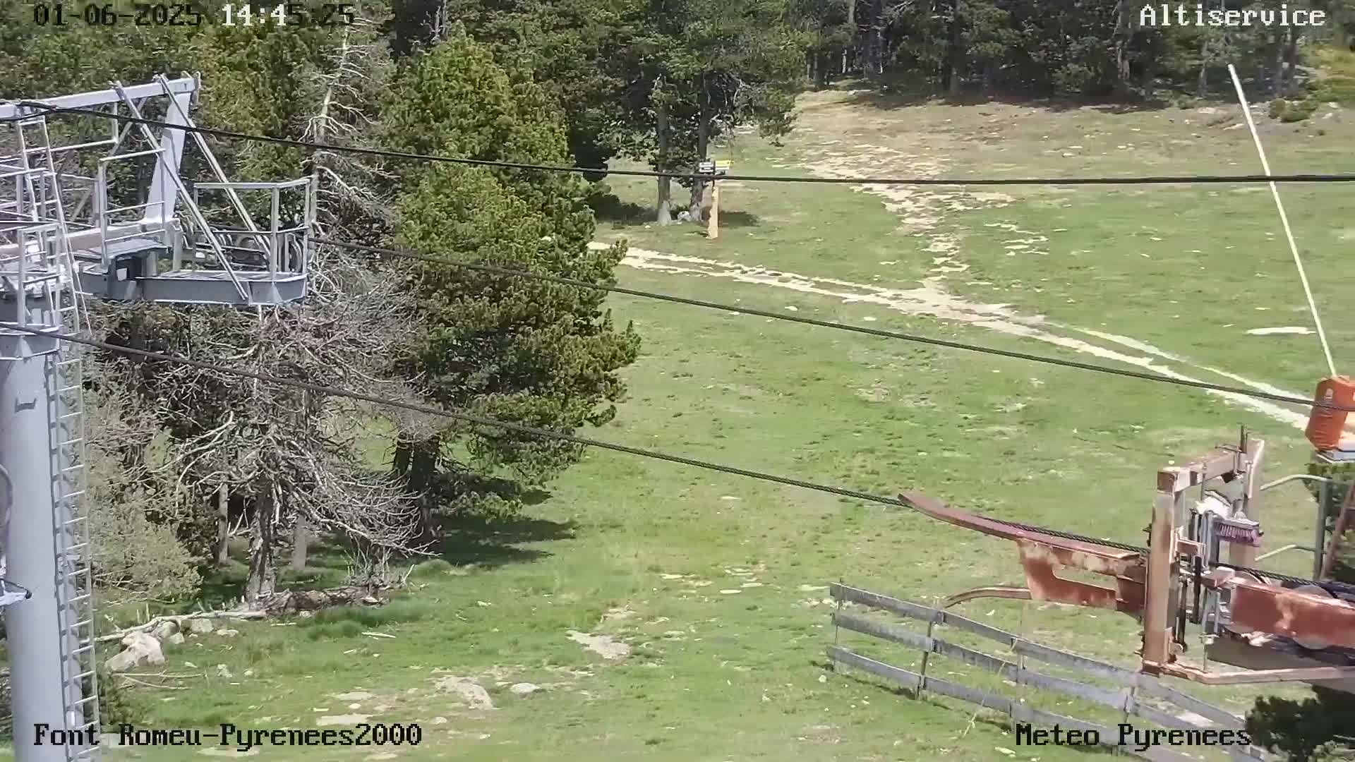 A chairlift mechanism is partially visible in the foreground, overlooking a grassy hillside with sparse trees and dirt paths under a sunny sky.