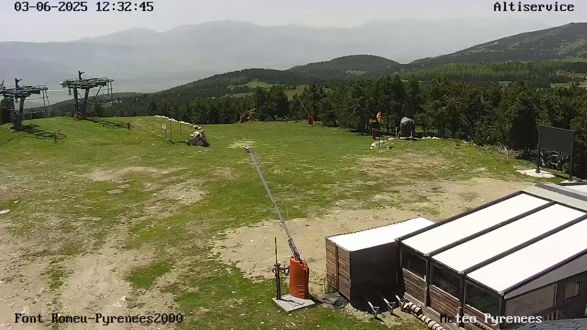 A mostly sunny, hazy view of a grassy mountaintop area with two ski lifts, a small building, and distant mountains.