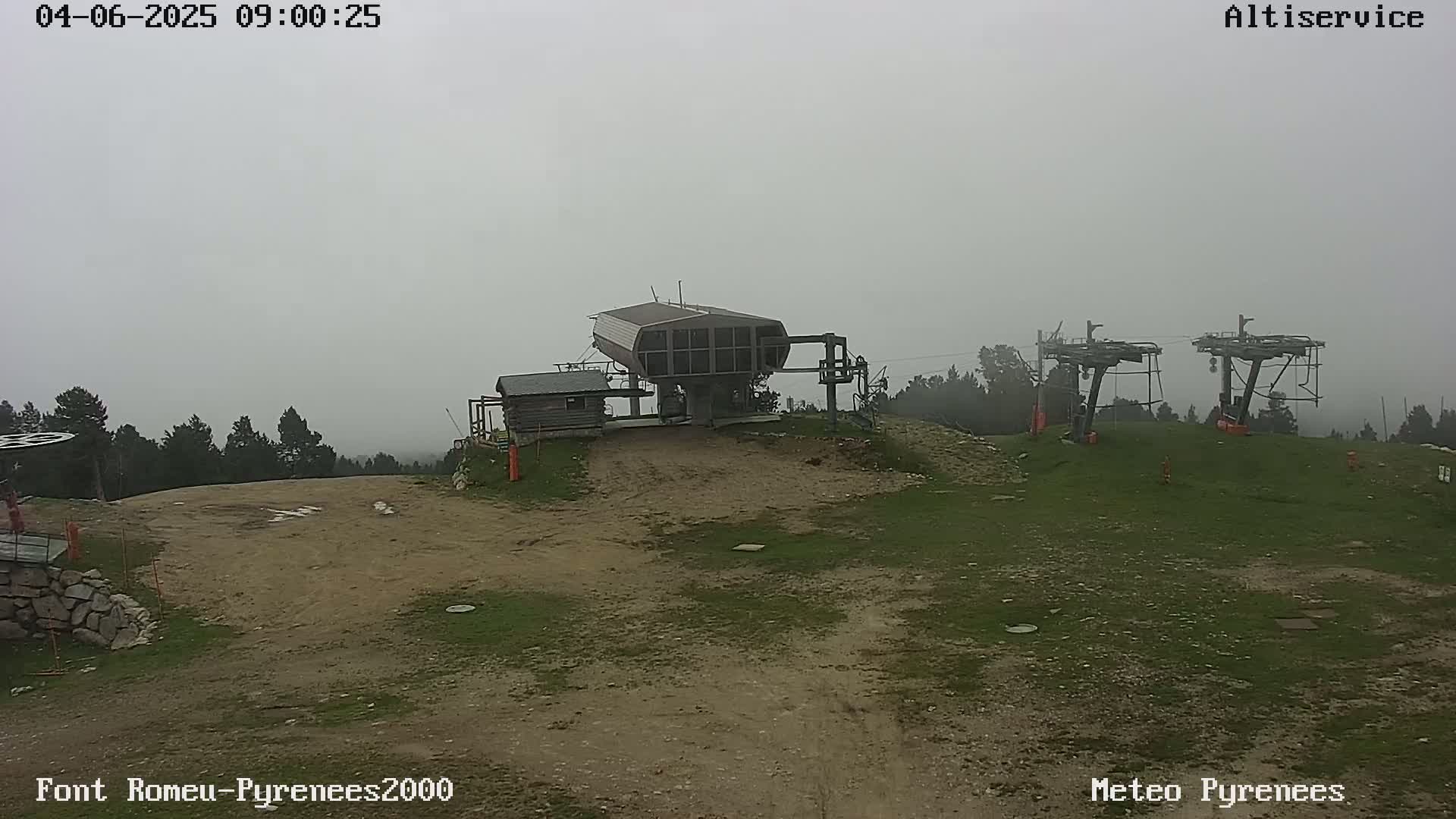 A foggy mountaintop view shows a ski lift terminal building and two lift towers on a grassy area with a dirt path, surrounded by a forest in the distance.