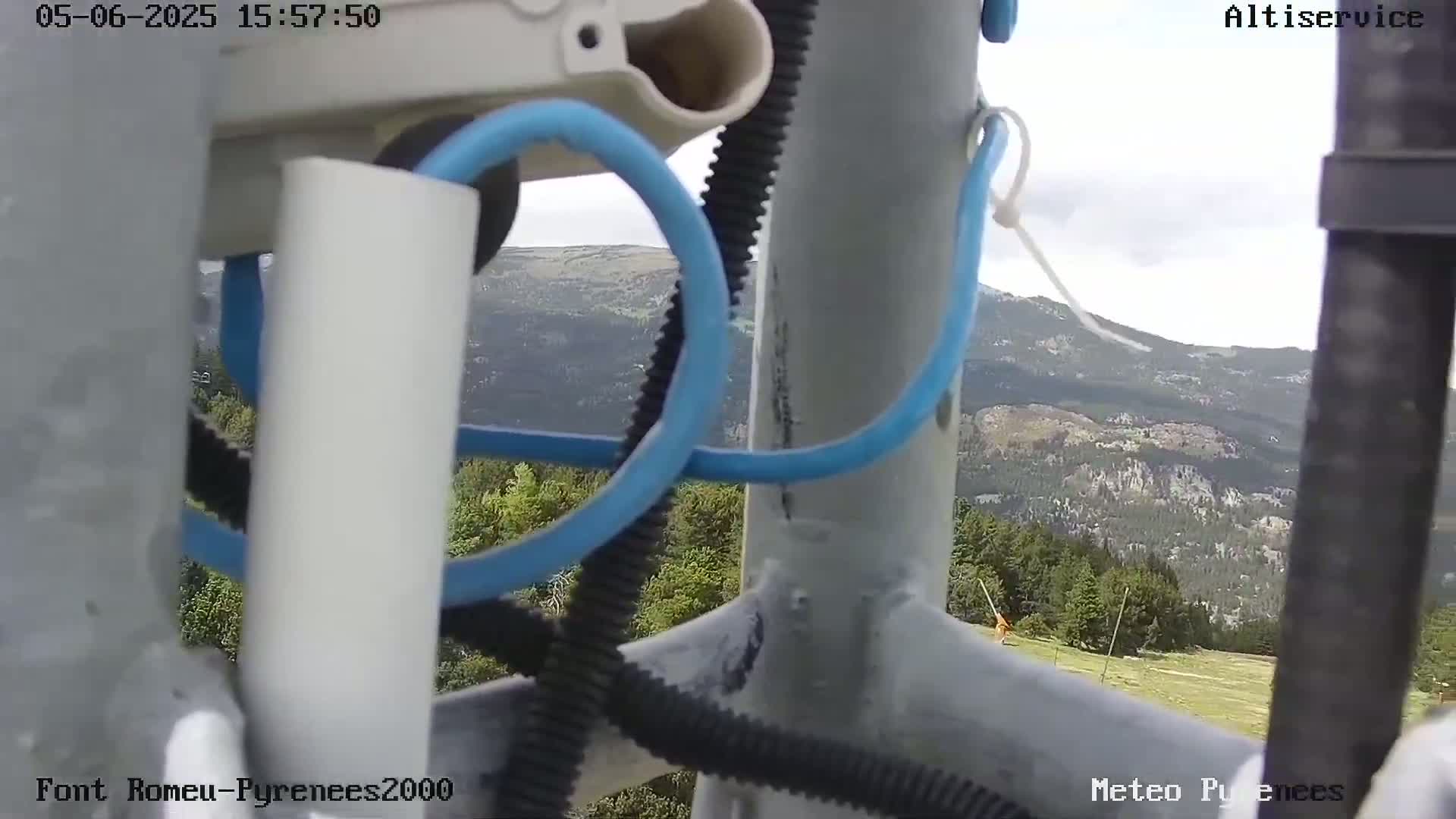 A close-up view of wires and metal poles with a mountainous landscape visible in the background under partly cloudy skies.