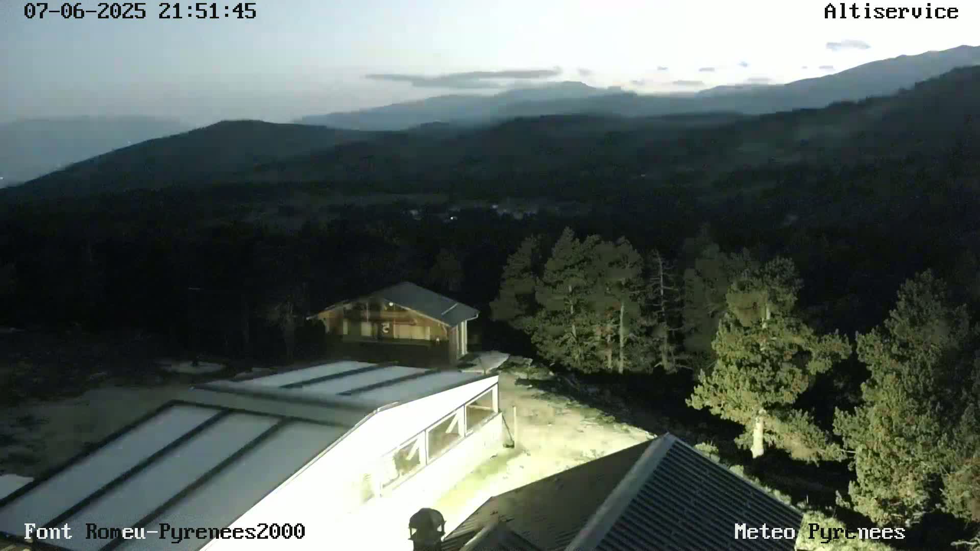 An elevated view at dusk shows several buildings nestled among dark green pine trees, with distant, hazy mountains in the background under a mostly clear sky.
