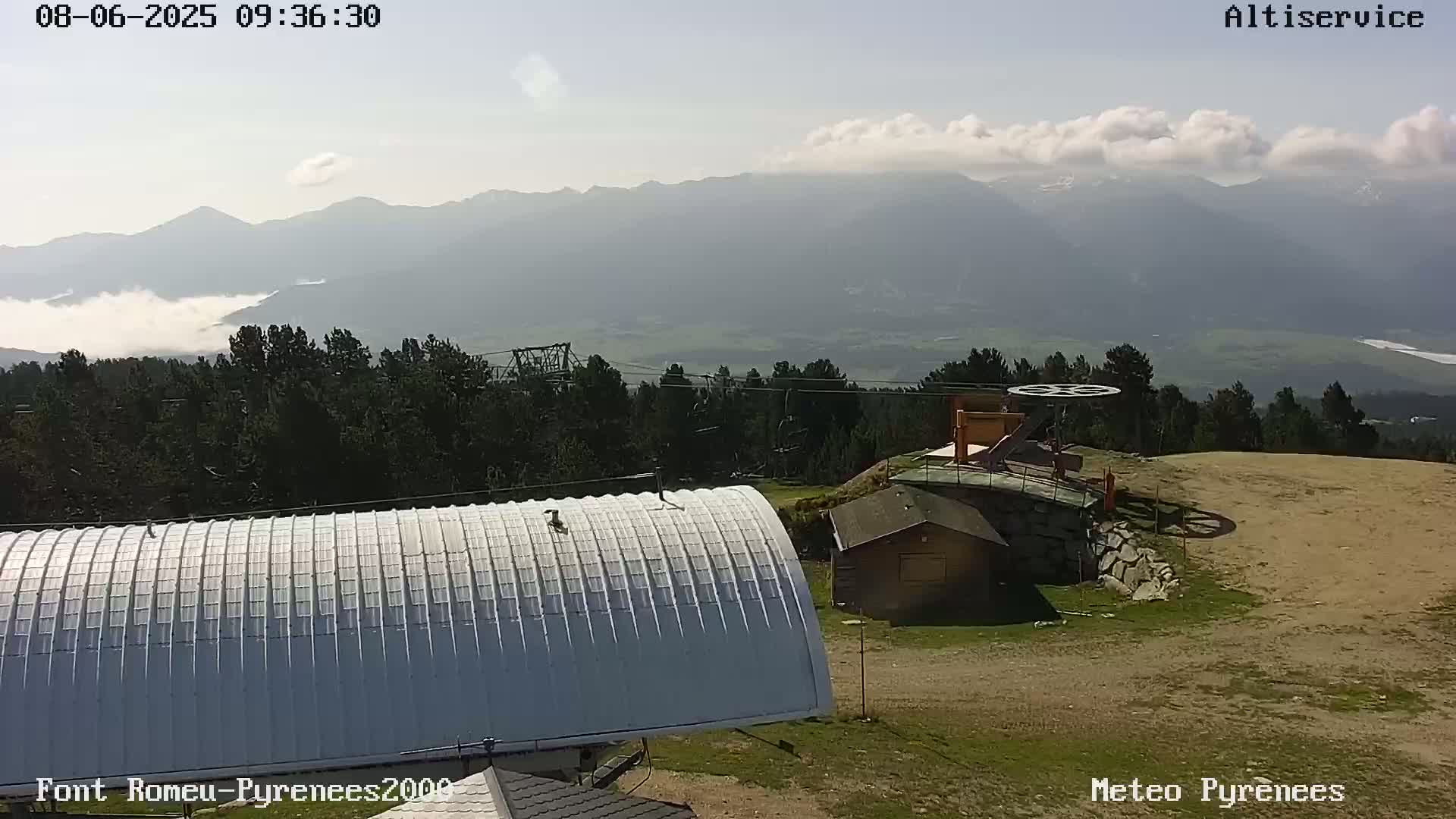 A mountaintop view on a partly cloudy day shows a curved metal roof, a small building, a ski lift mechanism, and a valley filled with low-hanging clouds and distant mountains.
