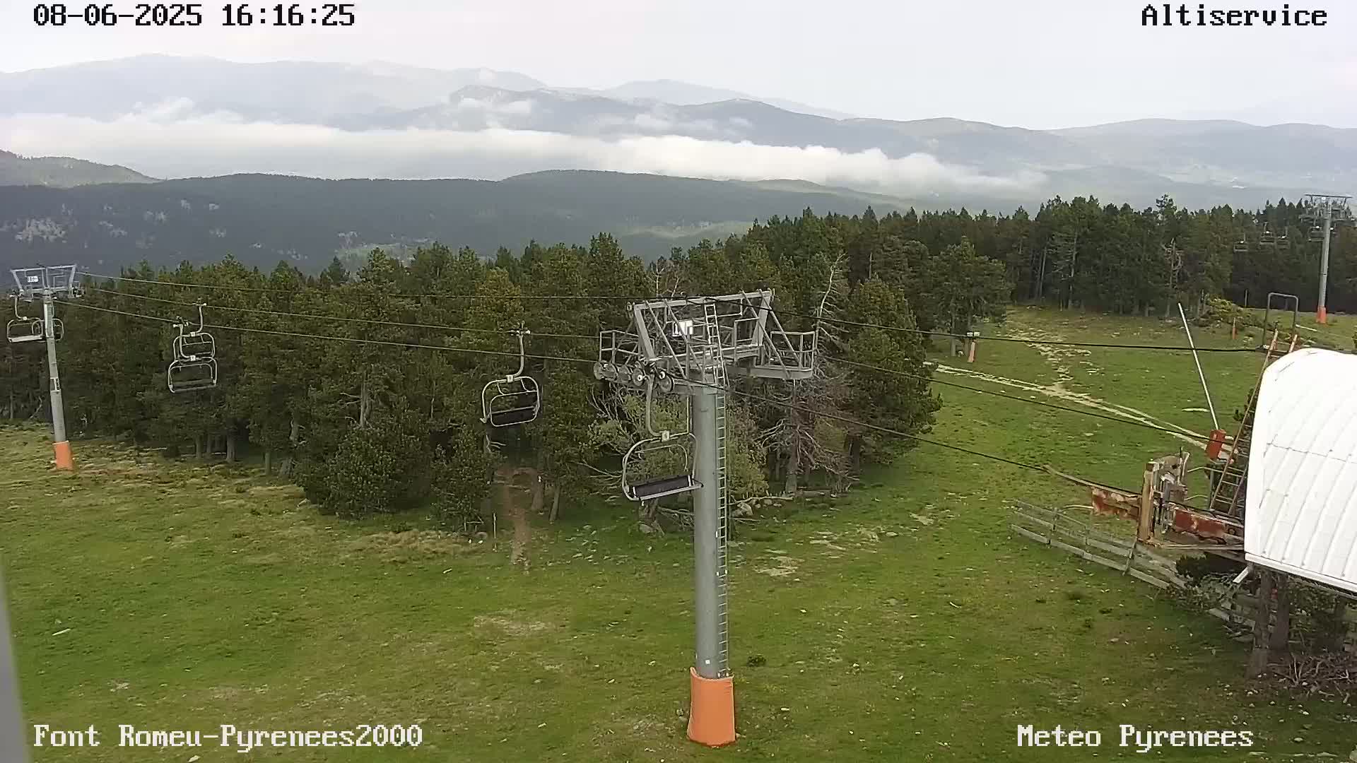 A chairlift stands in a grassy area amongst a pine forest, with a partly cloudy sky and mountains in the background.
