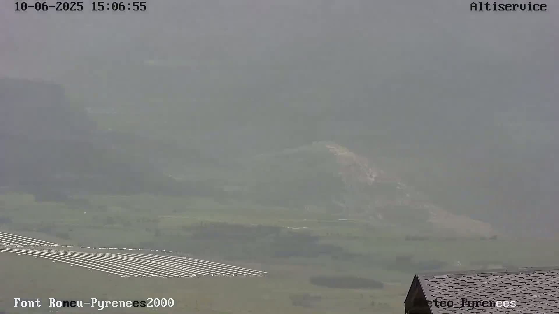 A hazy, foggy view shows a distant hillside, a section of what appears to be farmland with rows of plants, and a portion of a dark-roofed building in the bottom right corner.