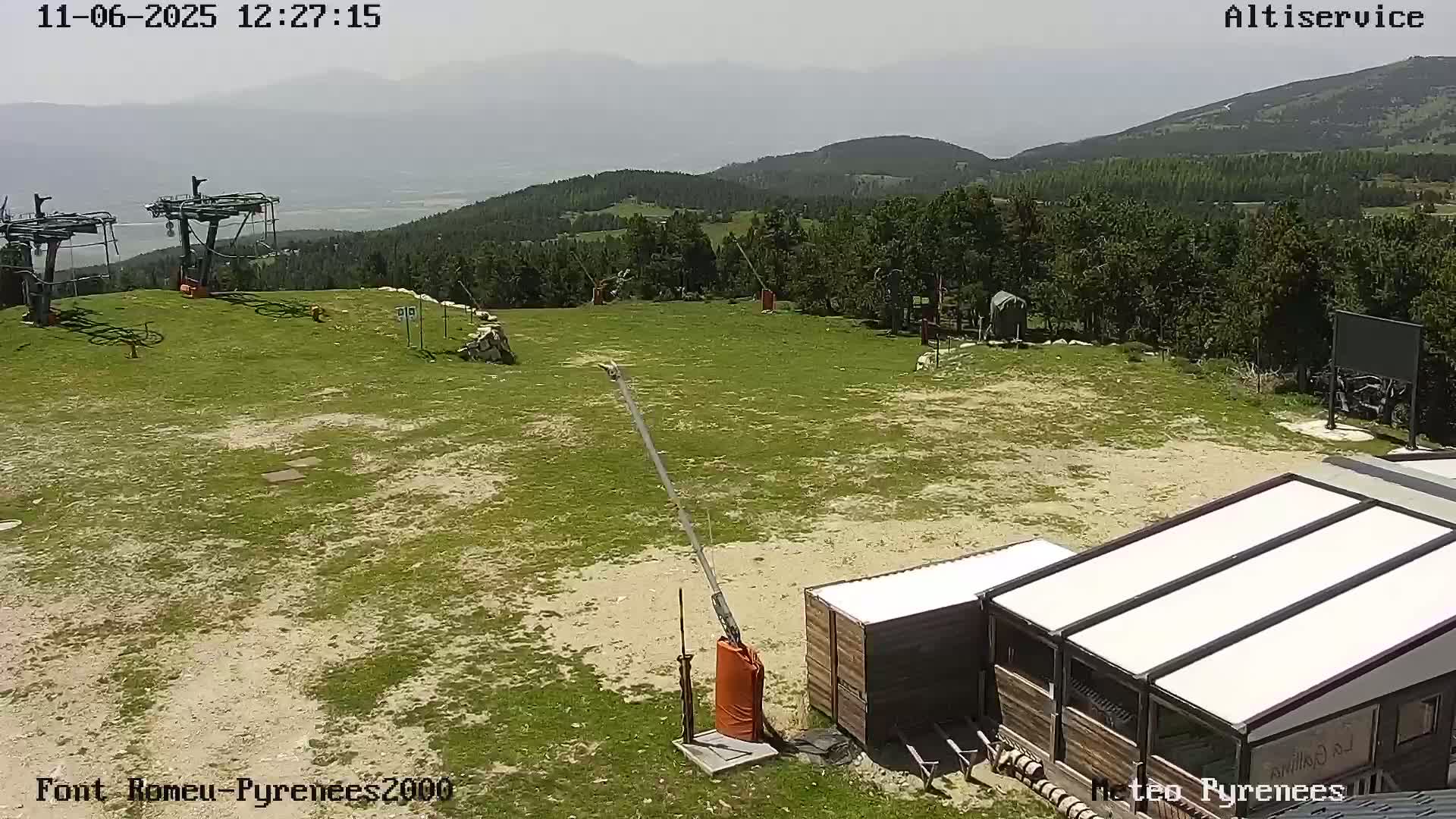 A mostly sunny day shows a grassy area with a ski lift, a small building, and a mountain view in the background.