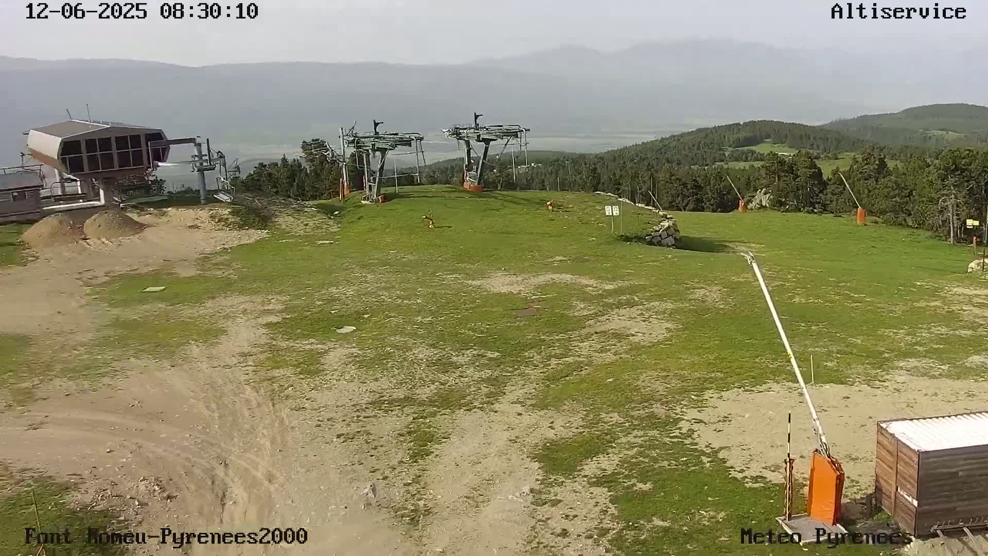A mostly clear day reveals two chairlift stations atop a grassy hill with distant mountains visible under a hazy sky.