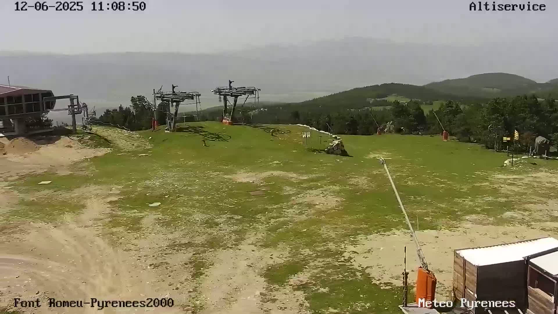 A mostly sunny, hazy day reveals a grassy ski lift area with two chairlifts and distant mountains.