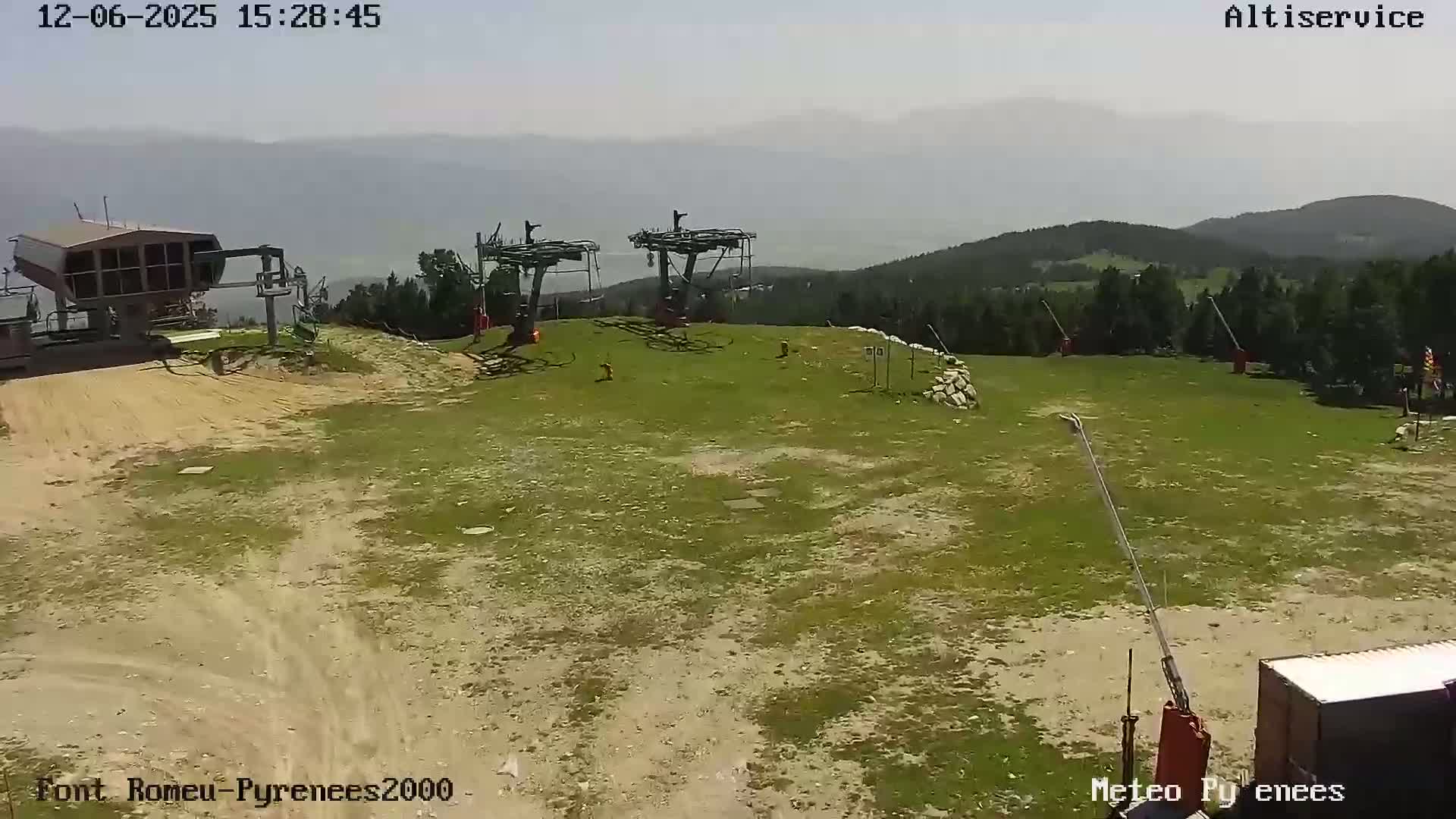 A mostly sunny day shows a mountaintop ski lift station with two chairlifts and a small building; the surrounding area is grassy with patches of dirt, and mountains are visible in the hazy distance.