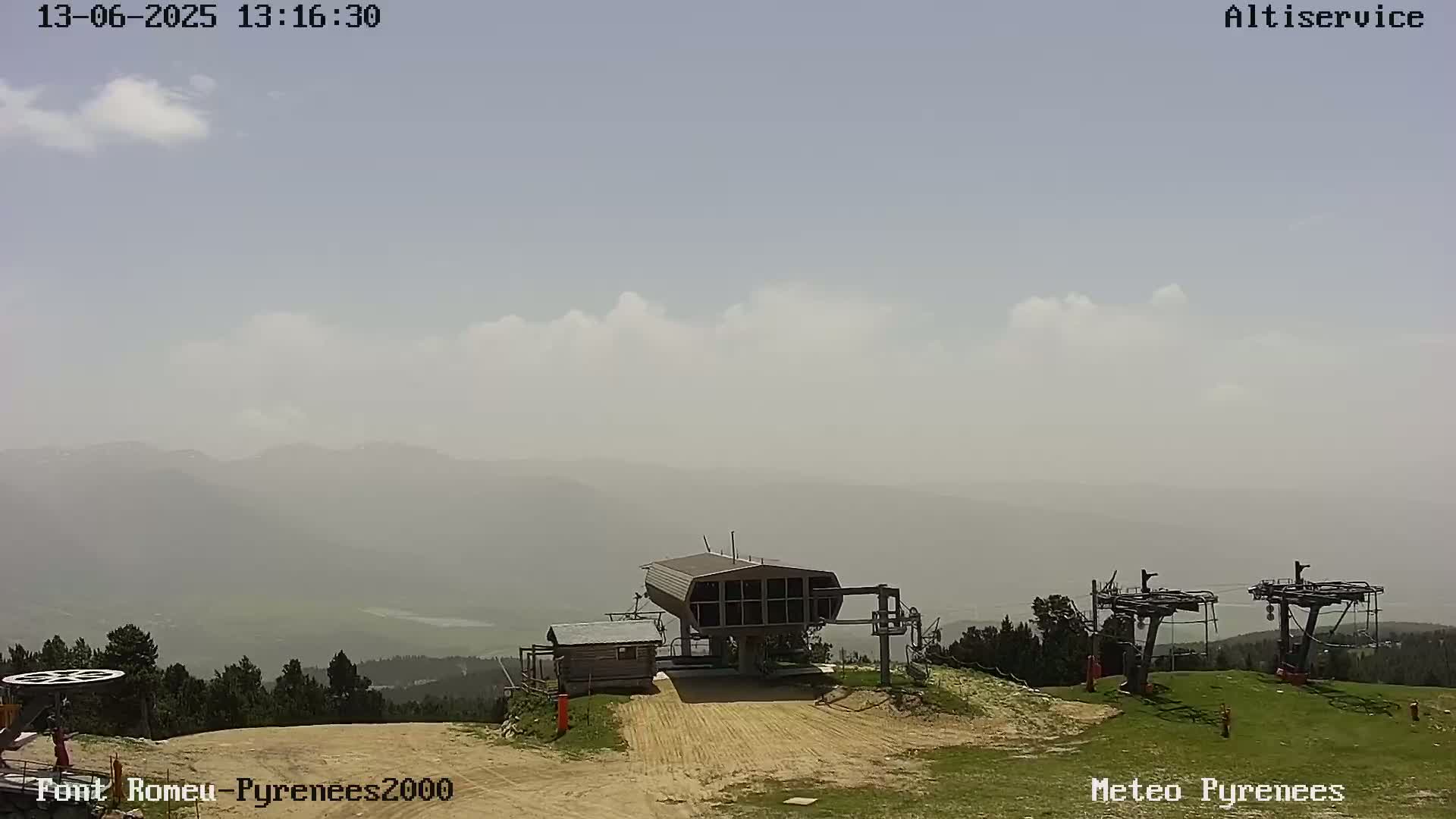 A hazy, sunny day reveals a mountaintop ski lift station with surrounding greenery and distant, hazy mountains.