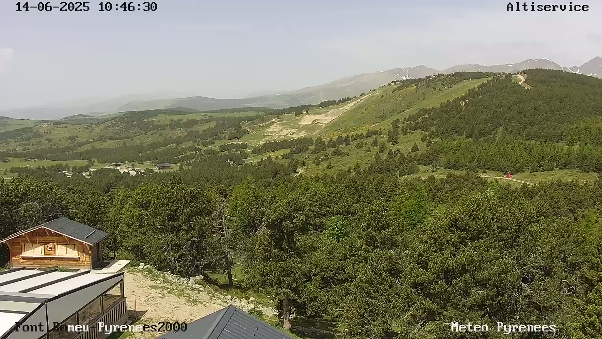 A sunny view of a mountainous landscape with green forests, a small building, and a cleared area on a hillside.