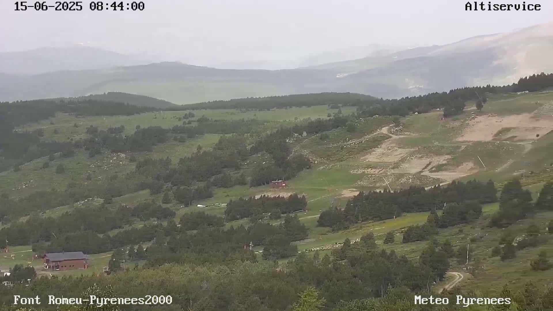 A hazy, overcast view shows a mountainous landscape with green hills, patches of bare earth, and some buildings, possibly a ski resort, nestled among the trees.