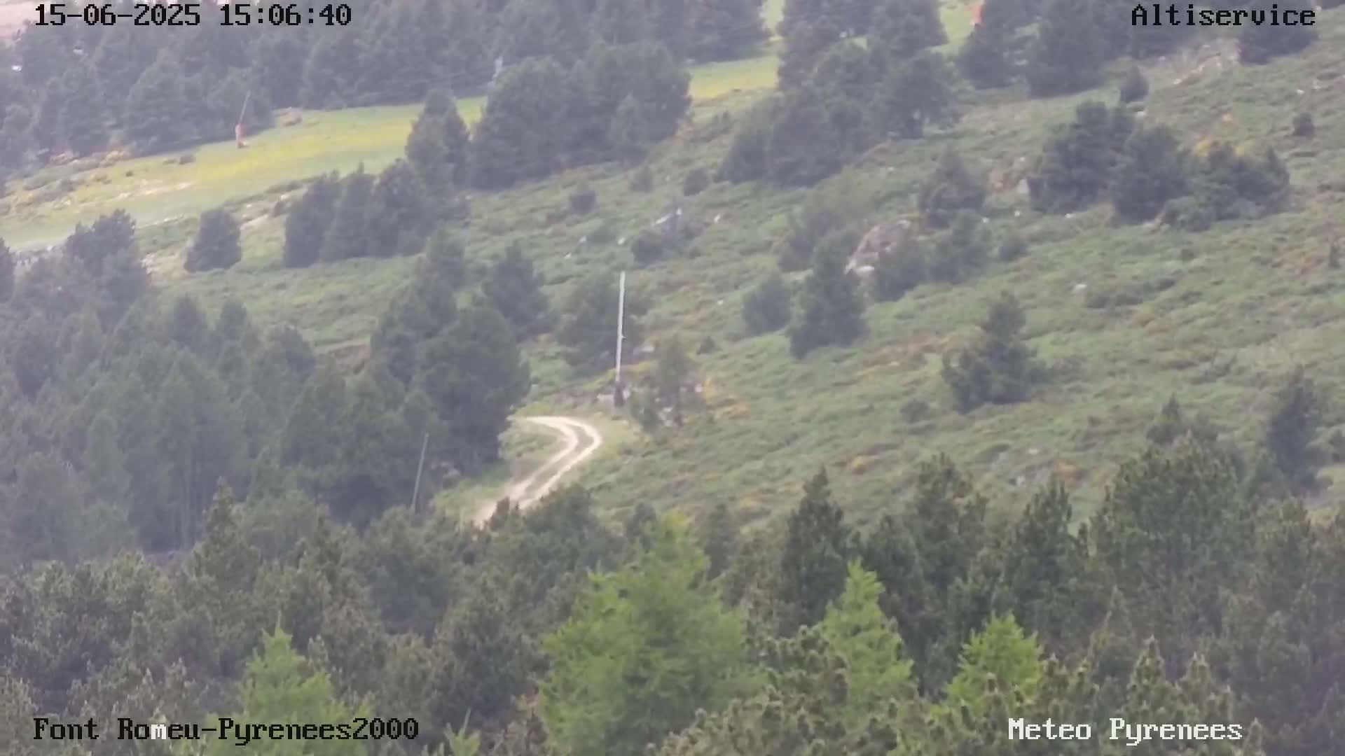 A hazy, high-angle view shows a winding dirt road through a mountainous landscape covered in green vegetation and coniferous trees.