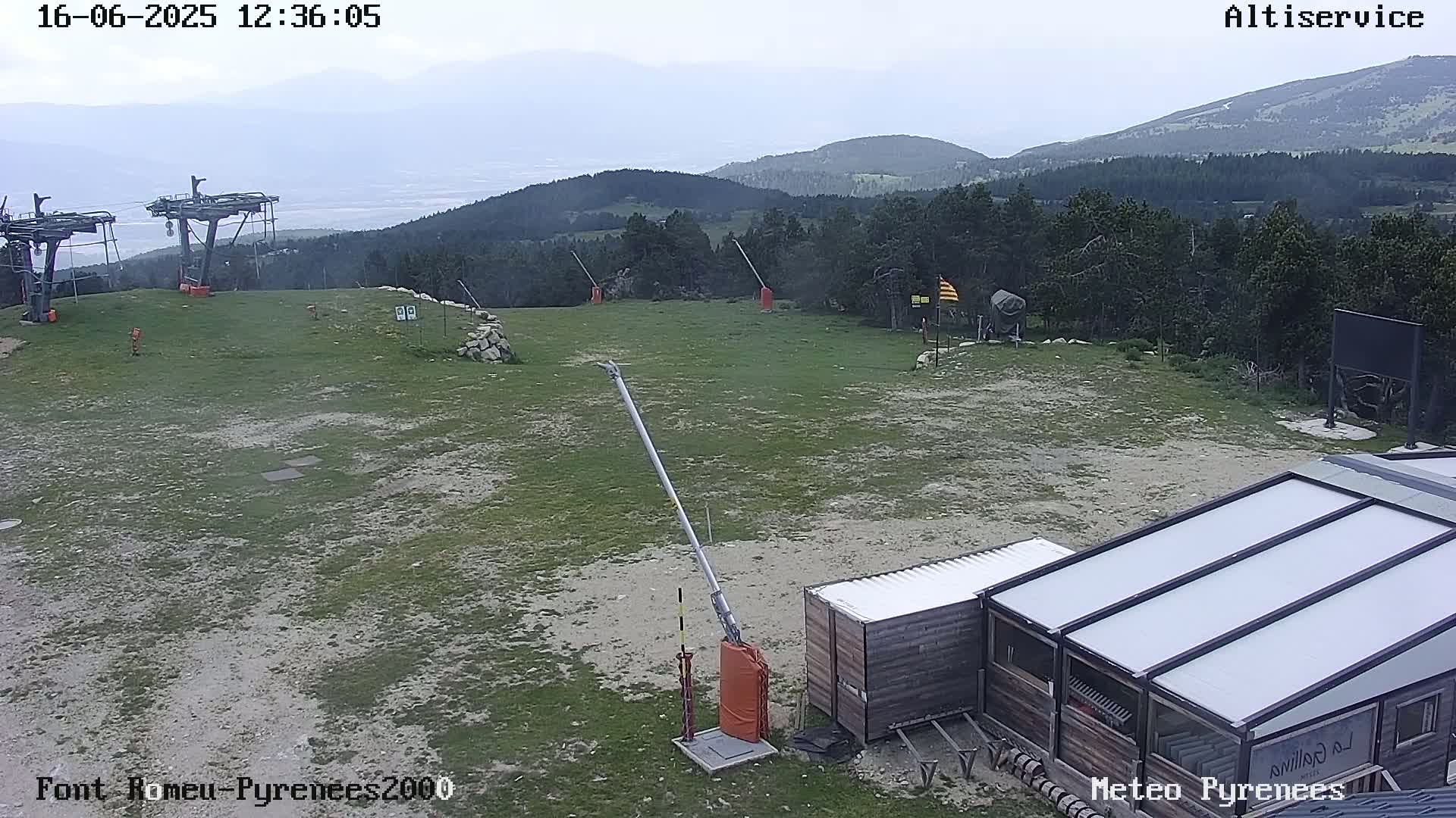 A mostly sunny, hazy day at a mountaintop ski lift area with grassy terrain, showing ski lift infrastructure and a small building.