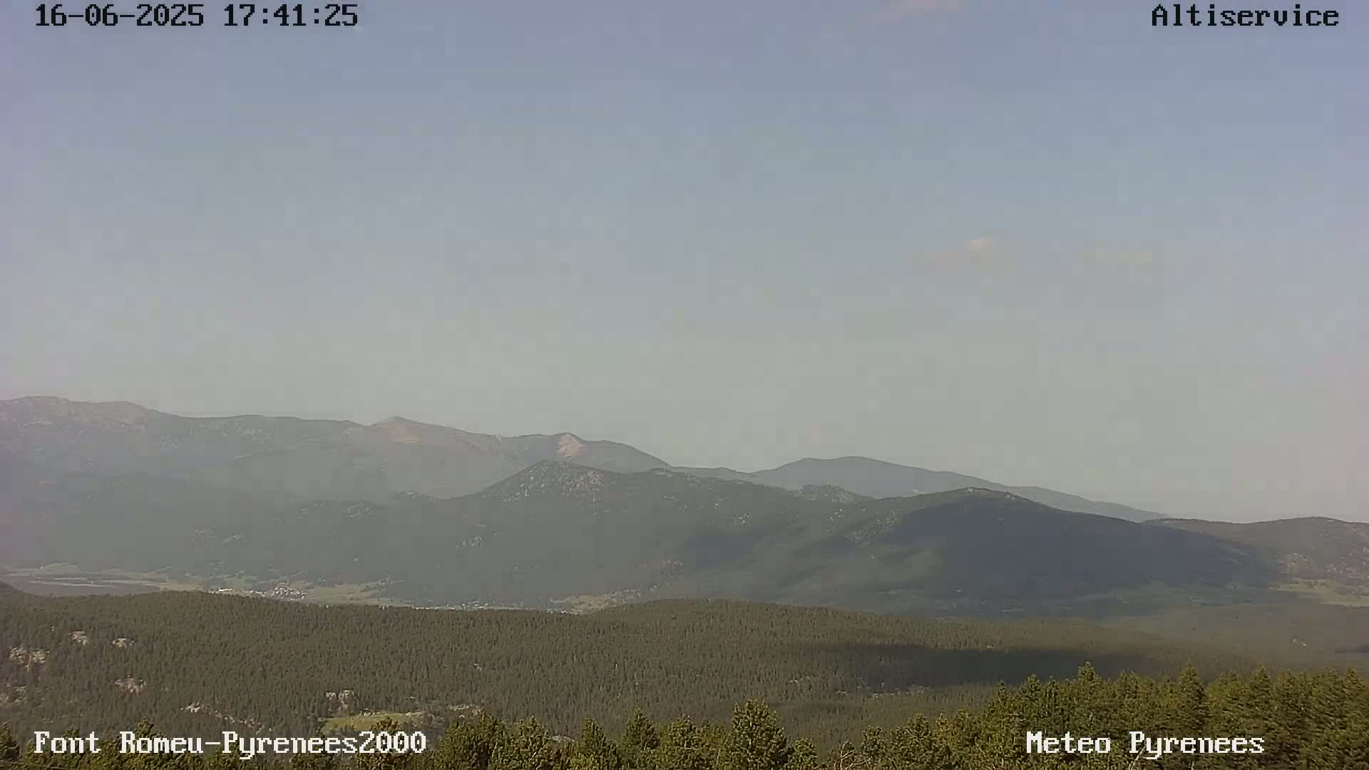 A hazy view shows a range of dark green mountains and a foreground of dark green evergreen trees under a light-blue, mostly clear sky.