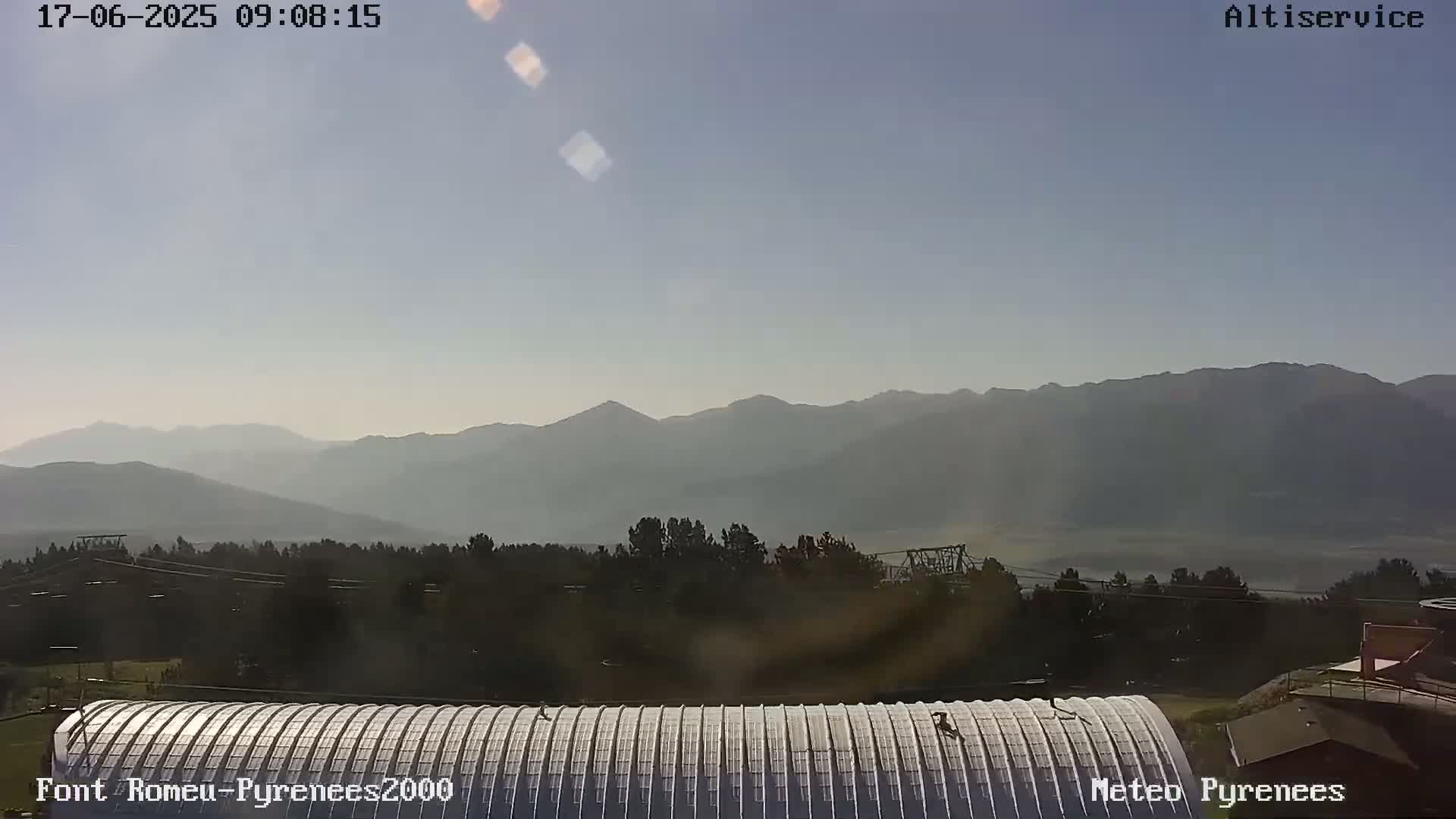 A hazy, sunny view of distant mountains is seen from above a corrugated metal roof, with some trees and buildings visible in the foreground.