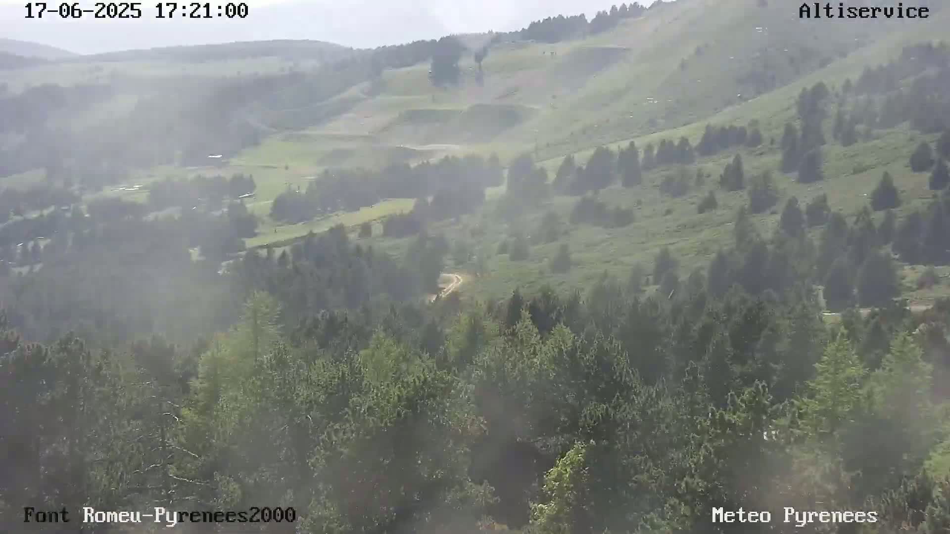 A hazy, long shot of a mountain landscape showing green hills, trees, and a dirt road, under a mostly clear sky.