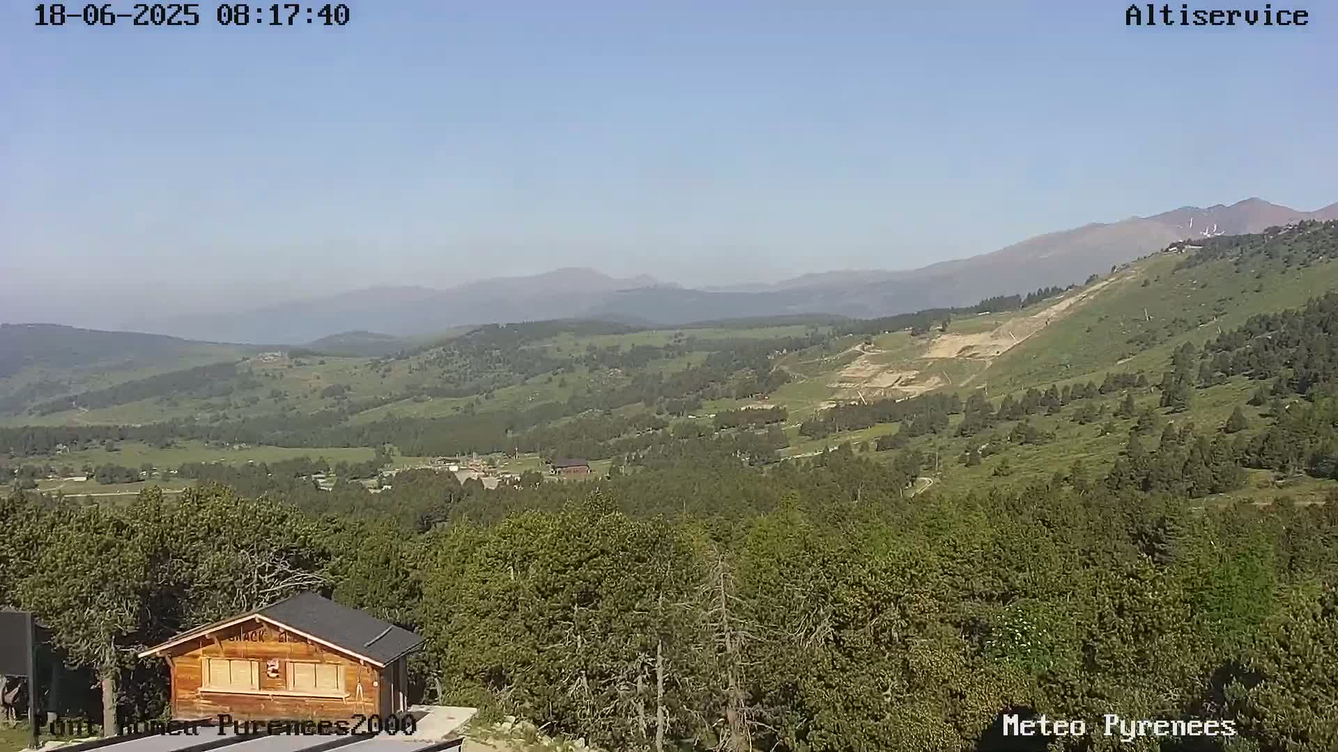 A panoramic view of a mountainous landscape under a clear blue sky shows a small wooden building in the foreground, a dense forest covering the middle ground, and rolling hills and distant mountains in the background.