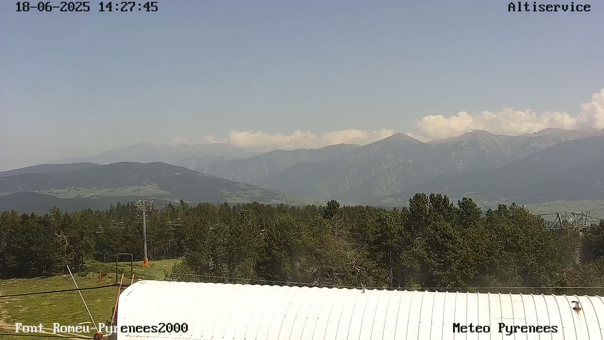 A partly cloudy sky overlooks a mountain range and a forest, with a white corrugated metal structure in the foreground.
