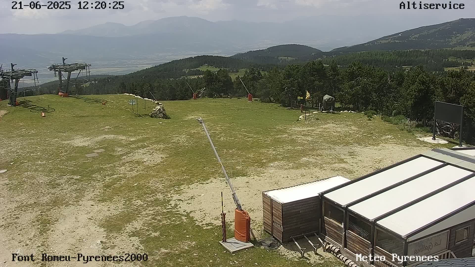 A mostly sunny, high-altitude view of a ski lift area with grassy terrain, a few small buildings, and distant mountains.