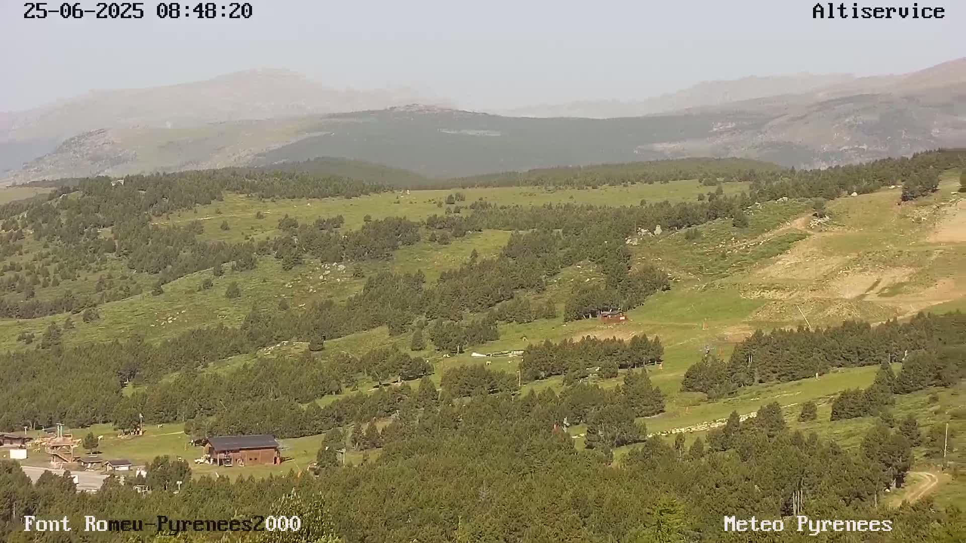 A hazy, sunny day reveals a mountainous landscape with green fields, scattered trees, and a few buildings in a valley.
