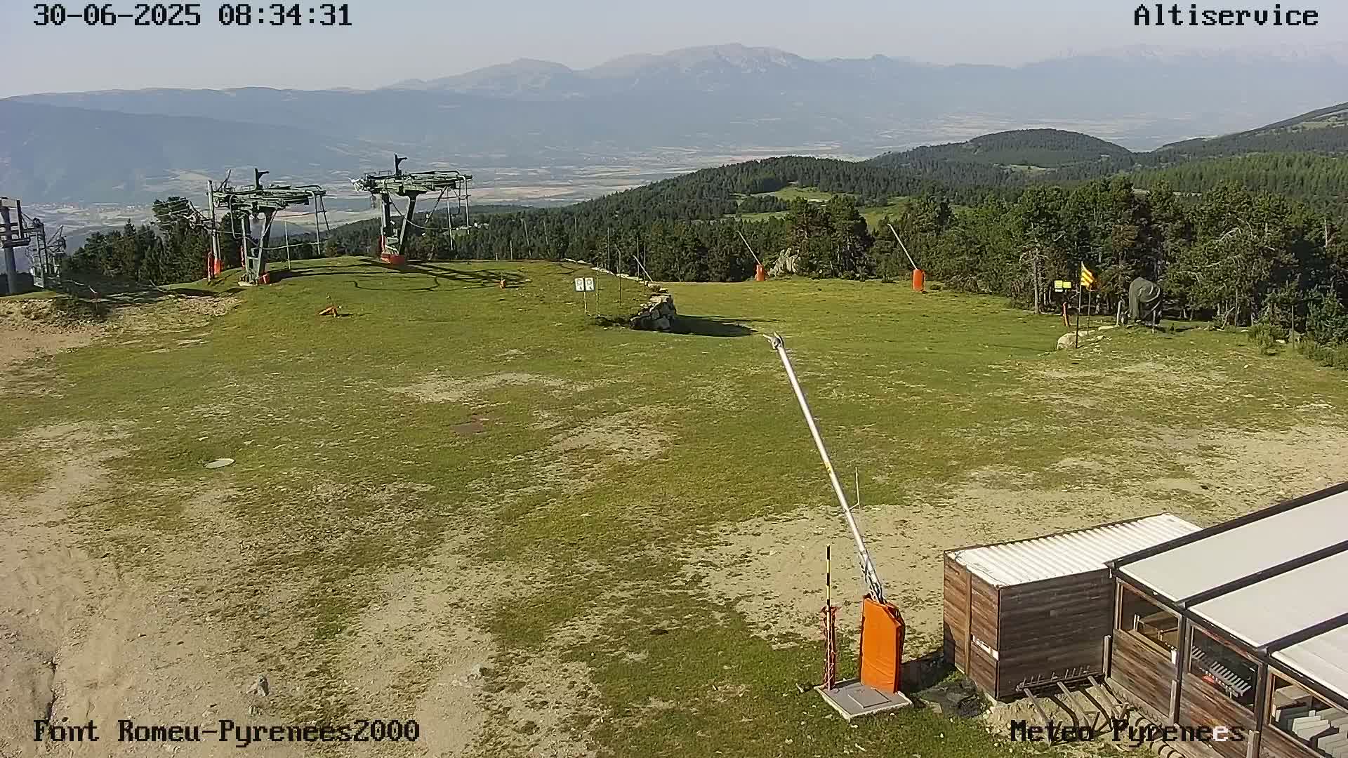 A grassy ski lift area with a small building and mountain range in the background under sunny conditions.