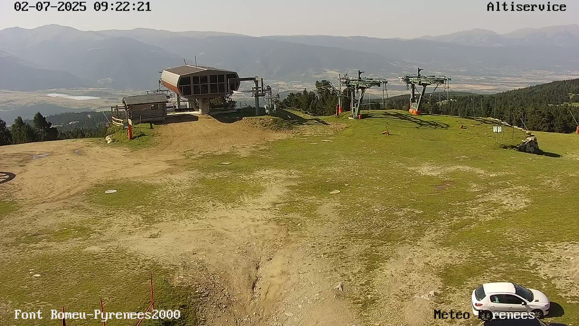 A mostly sunny mountaintop scene shows two chairlift stations, a small building, a dirt and grass area, and a single car parked nearby, with mountains visible in the background.